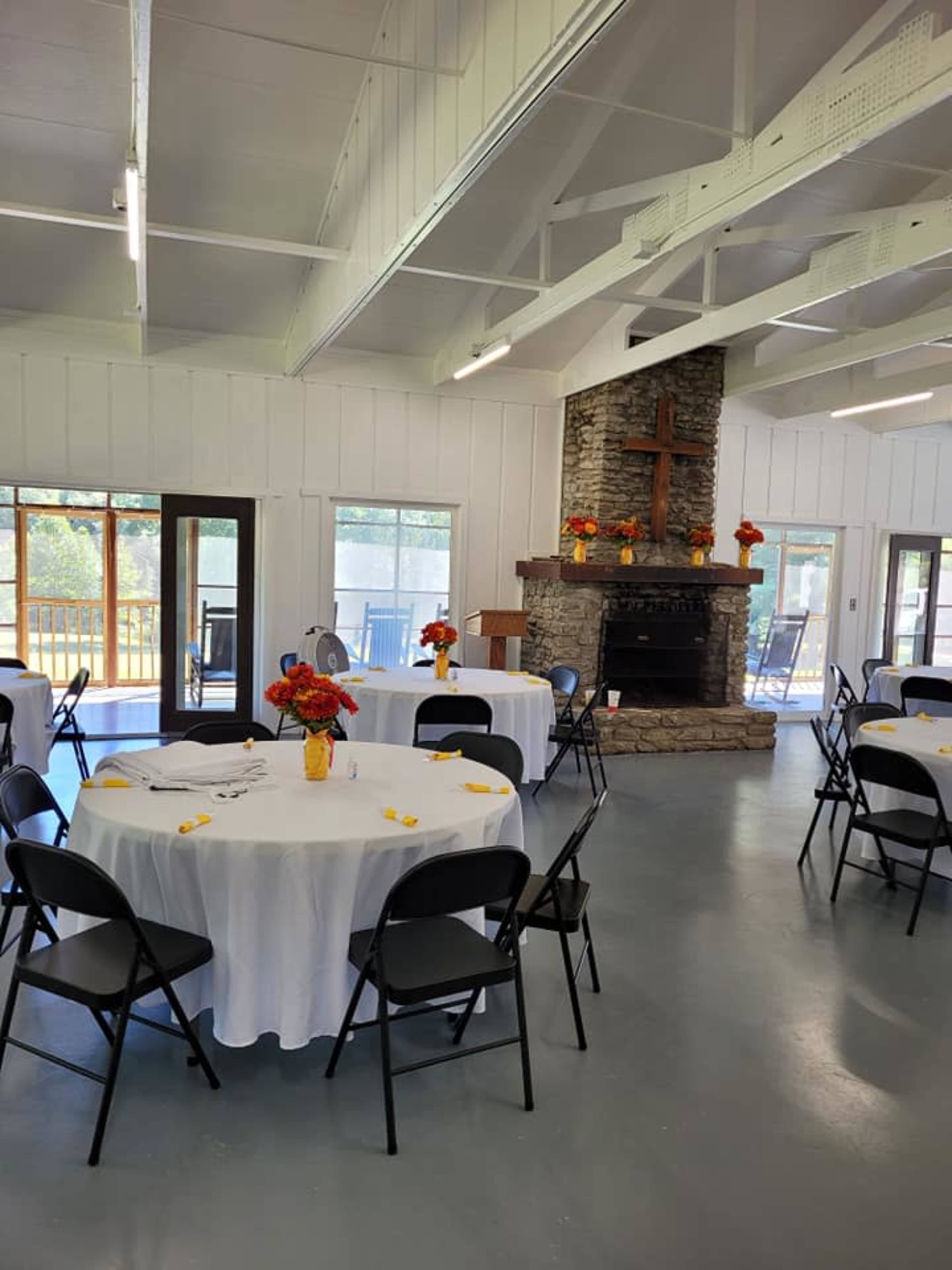 A spacious indoor setting with several round tables covered in white tablecloths and decorated with flower centerpieces, alongside a stone fireplace and a wooden cross.