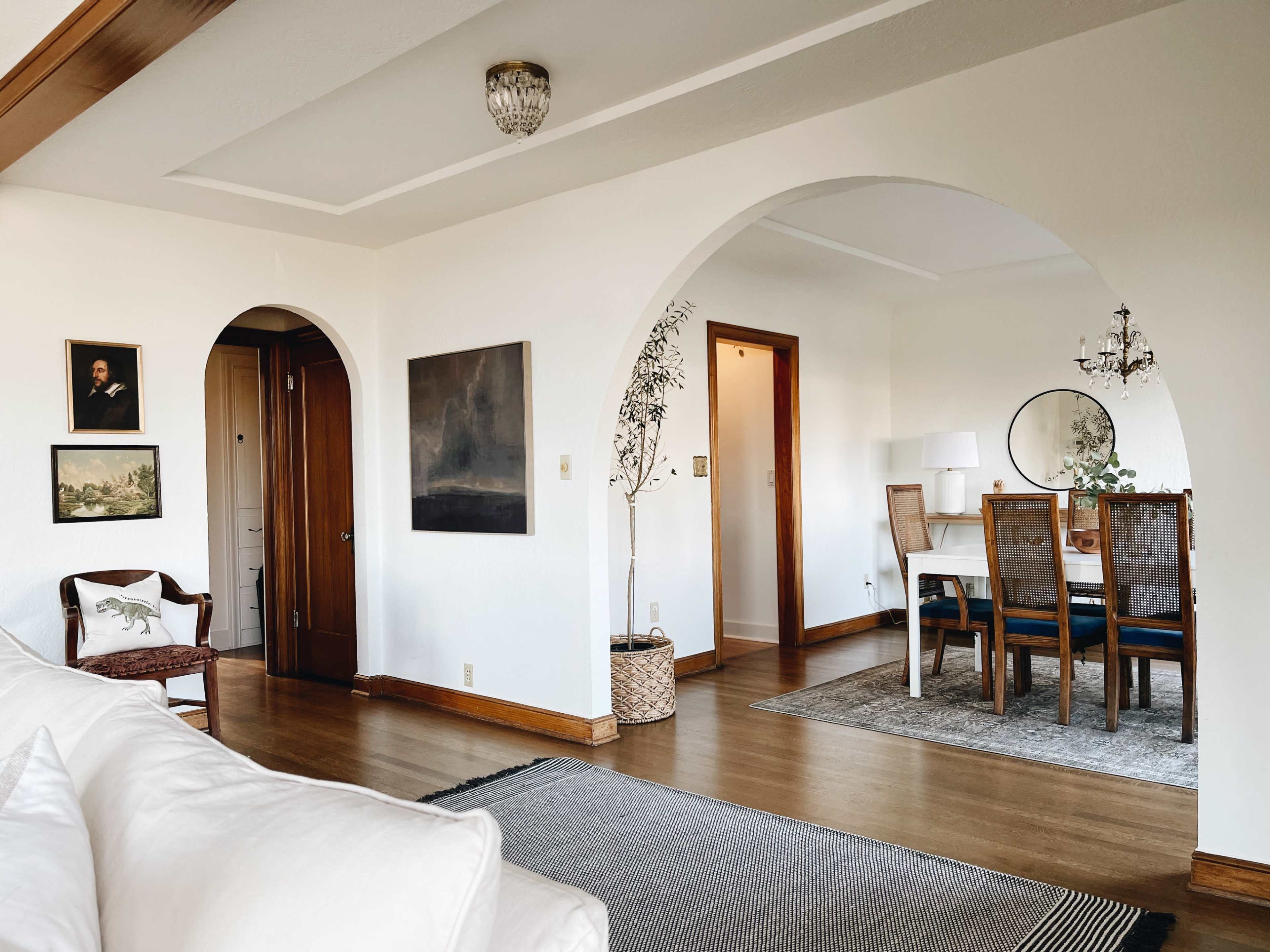 A spacious living area features an arched doorway leading to a dining space with a table and chairs, framed artwork on the walls, and a neutral color scheme.
