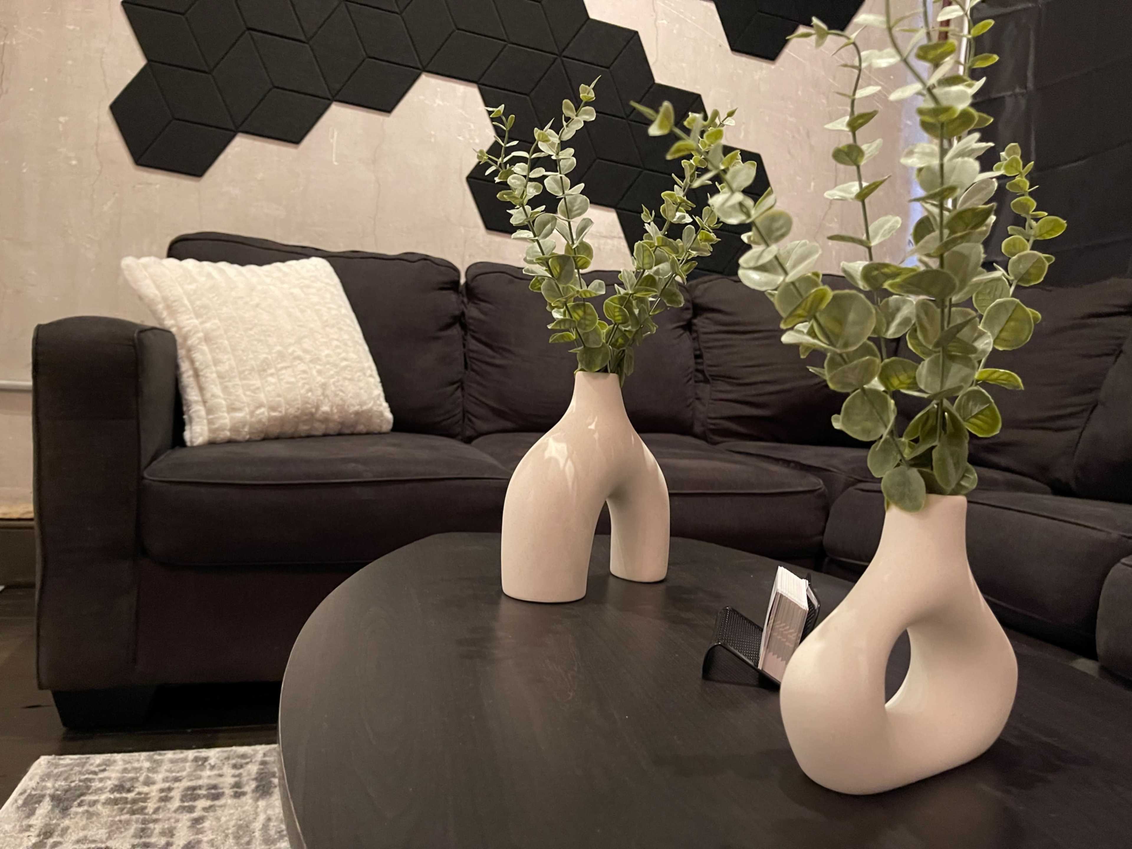 The image shows a modern living room scene featuring a black couch, a round dark table, and two white vases with greenery arranged on the table.
