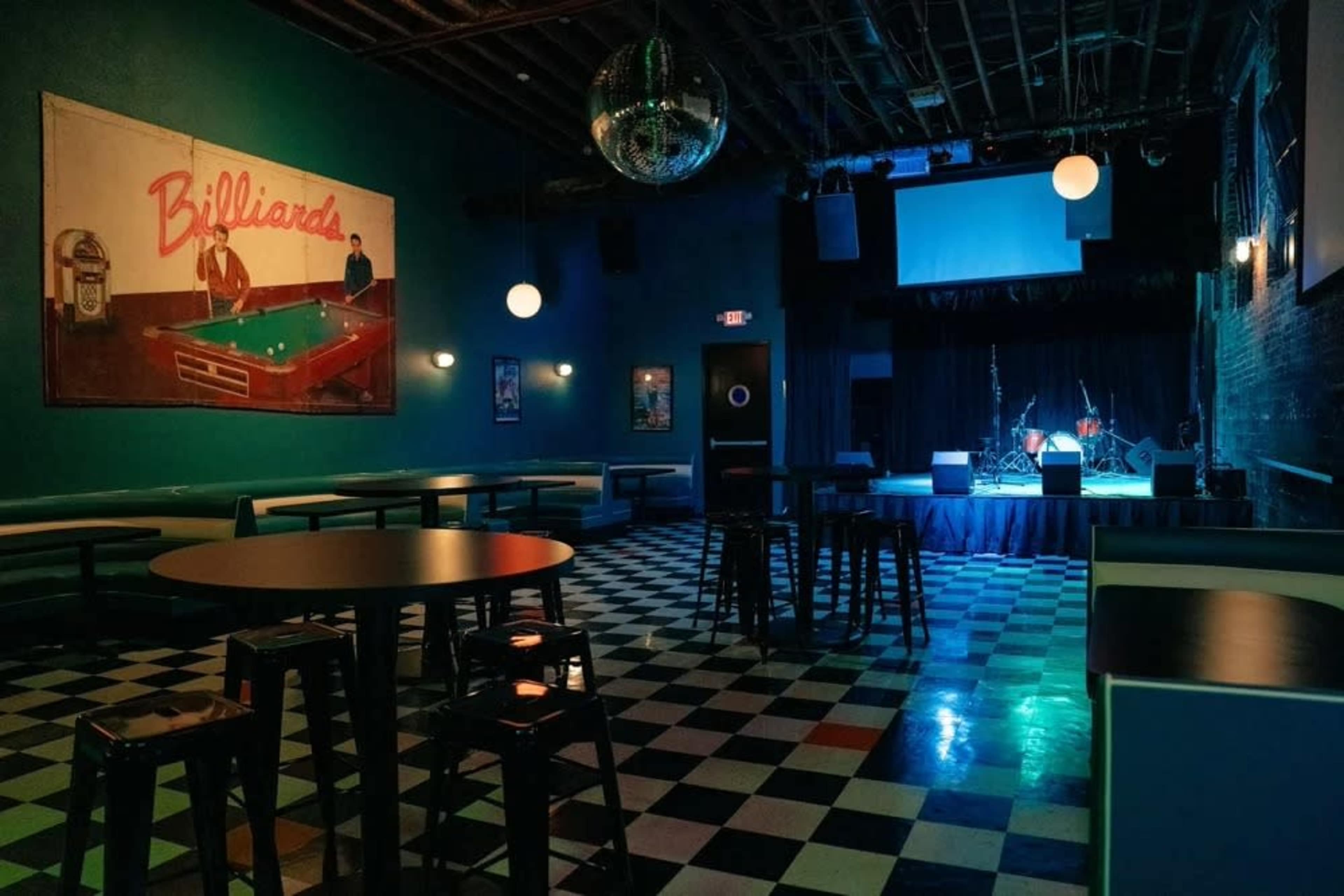 The image shows an empty venue with checkered flooring, a stage at the front, and a mural of a billiards scene on the wall.