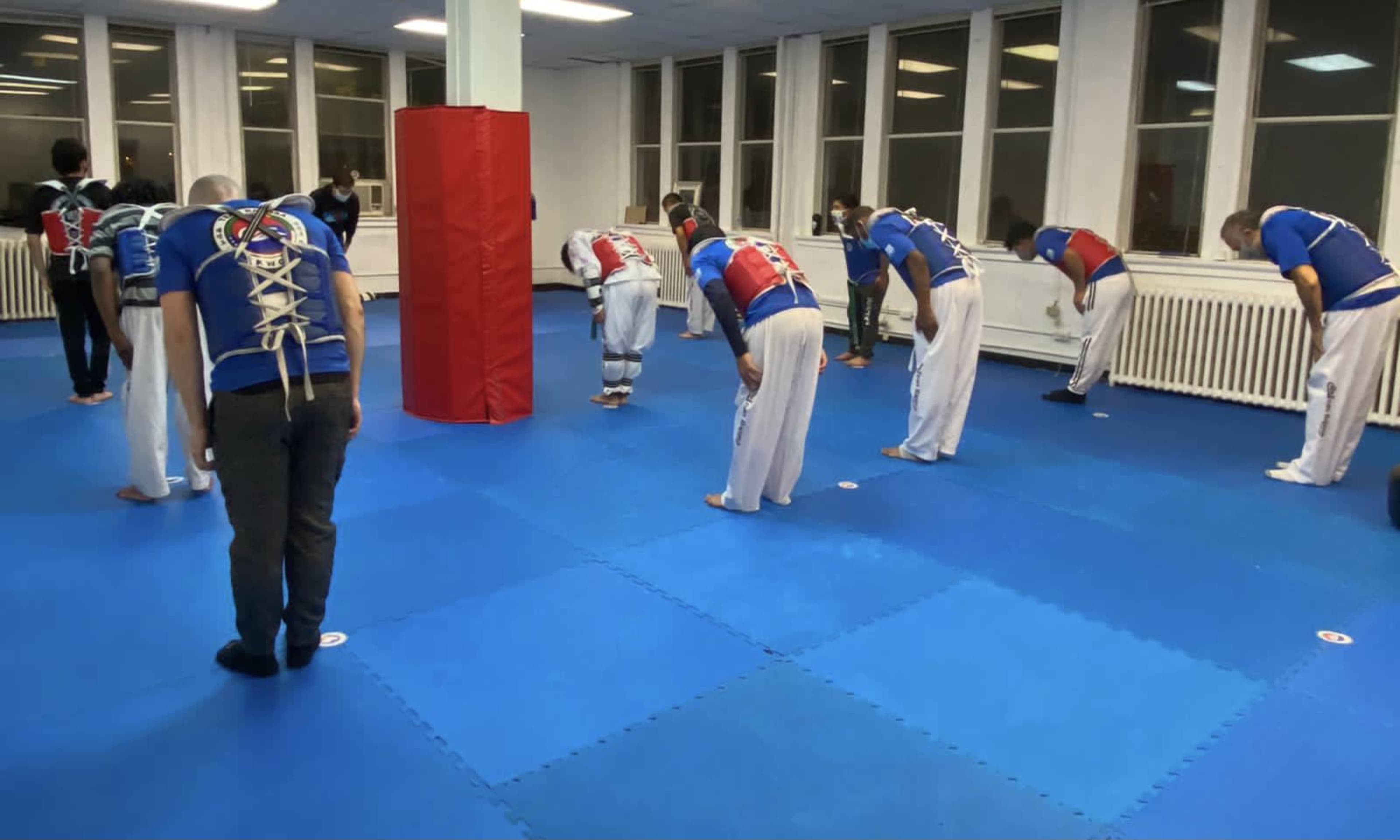 A group of individuals in martial arts uniforms are bowing in a training studio with blue mats and large windows.