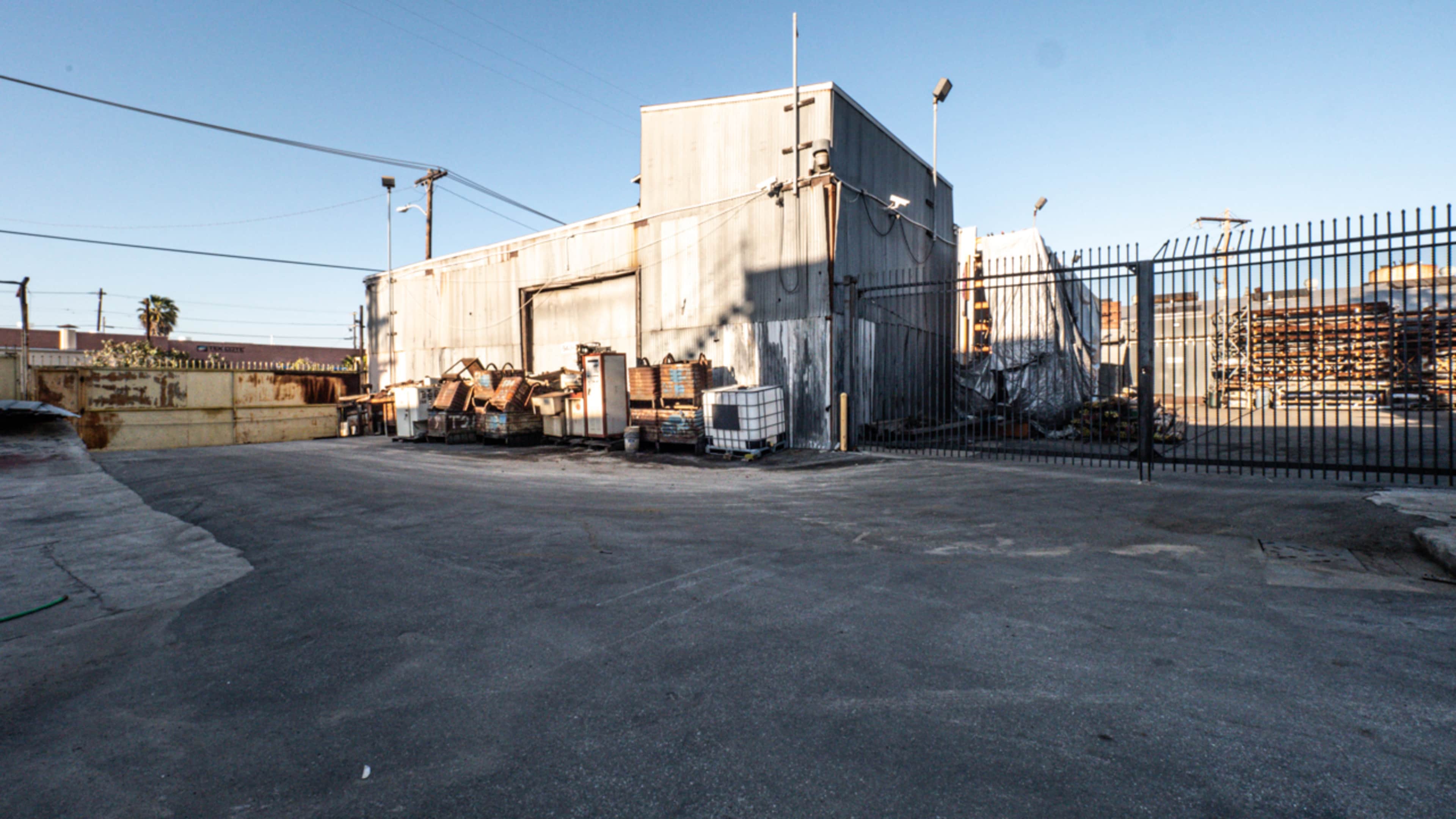 The image shows a barren concrete lot with a metal building and stacks of materials behind a gated fence.