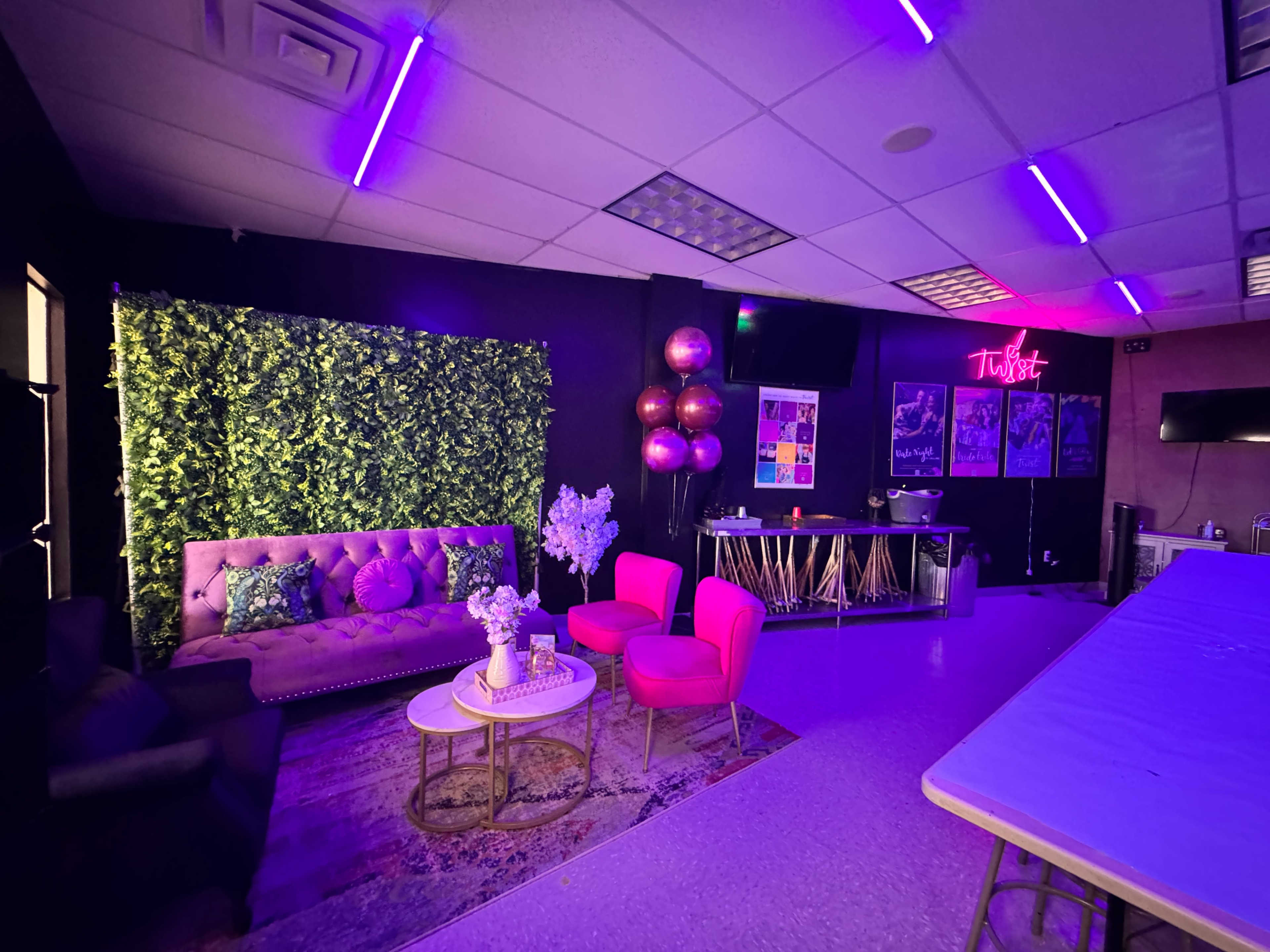 The interior features a lounge area with a purple sofa, pink chairs, and a decorative green wall, illuminated by purple lights.