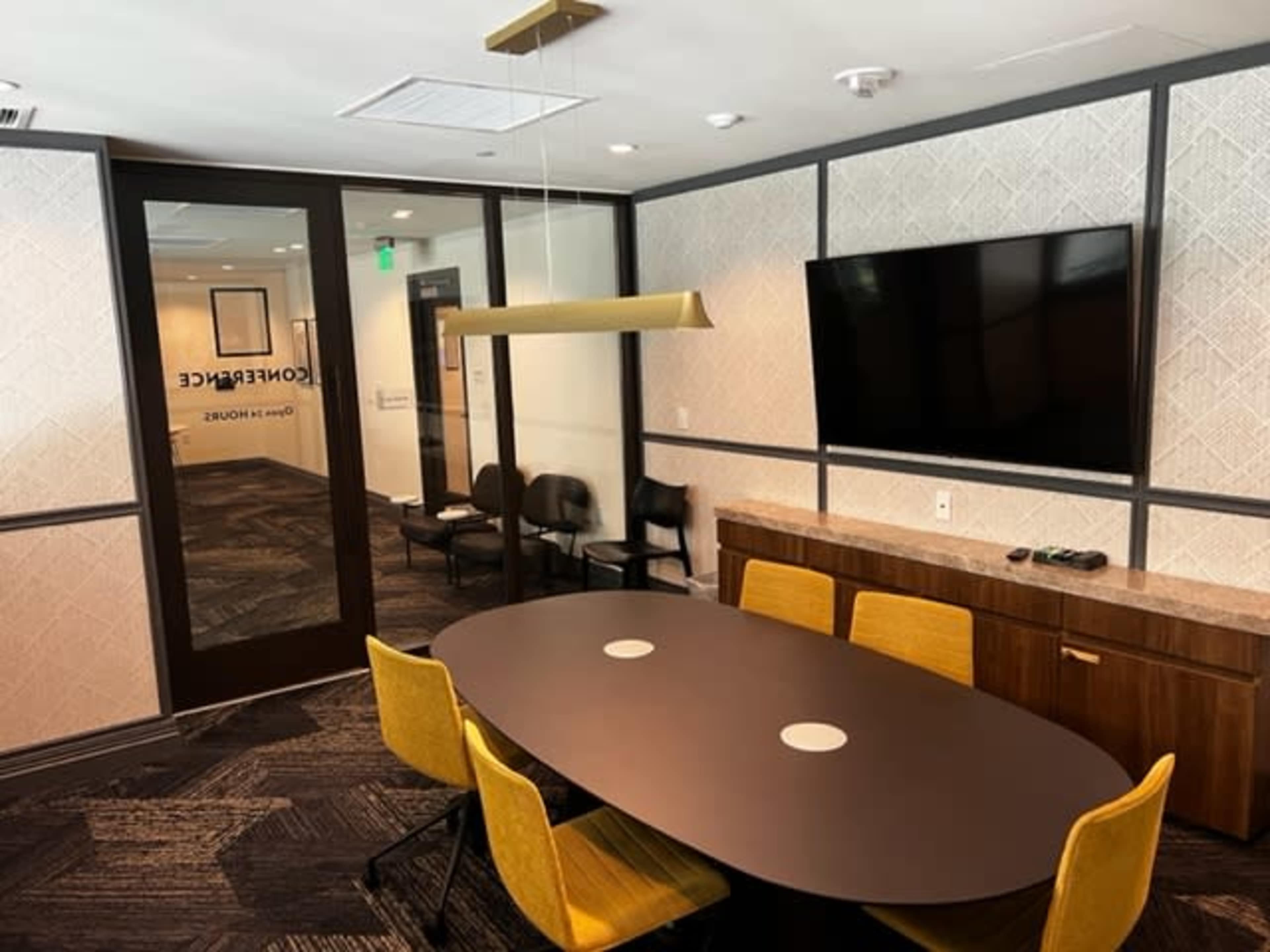 A modern conference room with a round table, yellow chairs, a television mounted on the wall, and a glass door labeled "Conference."