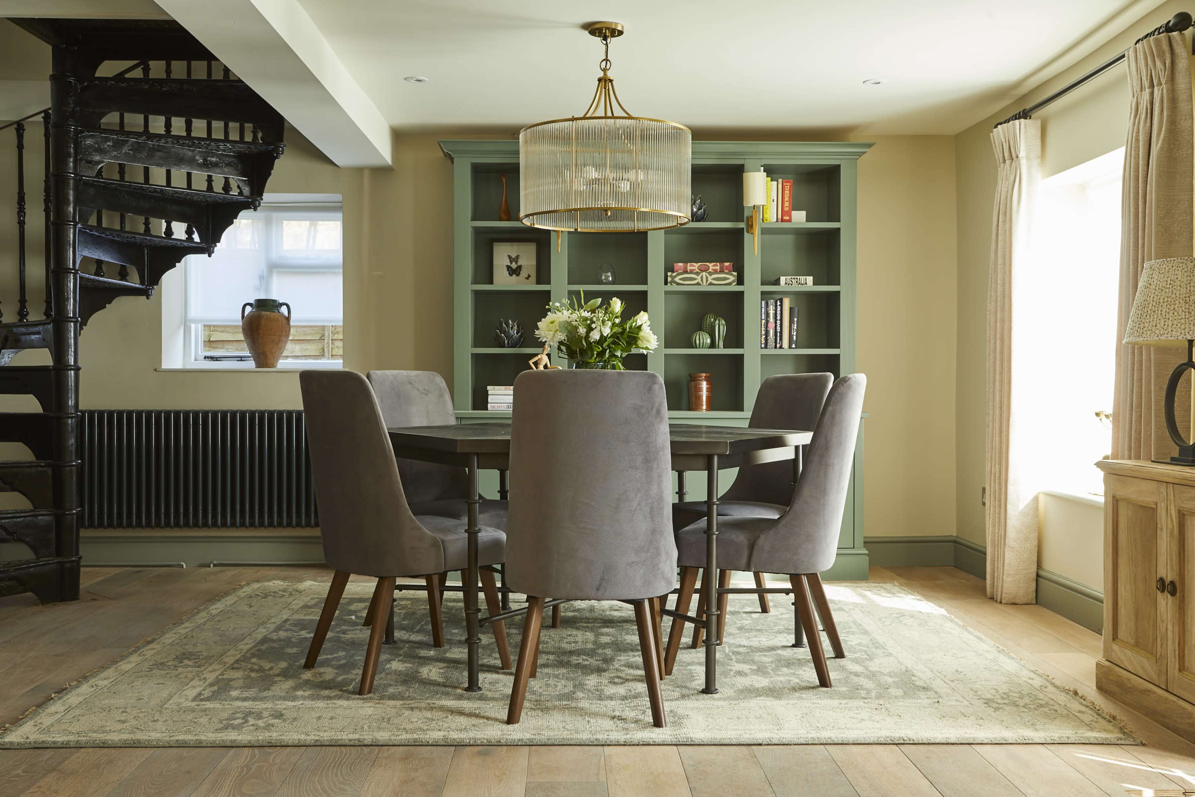 A dining area features a round table surrounded by six gray upholstered chairs, with a spiral staircase and built-in bookshelf in the background.