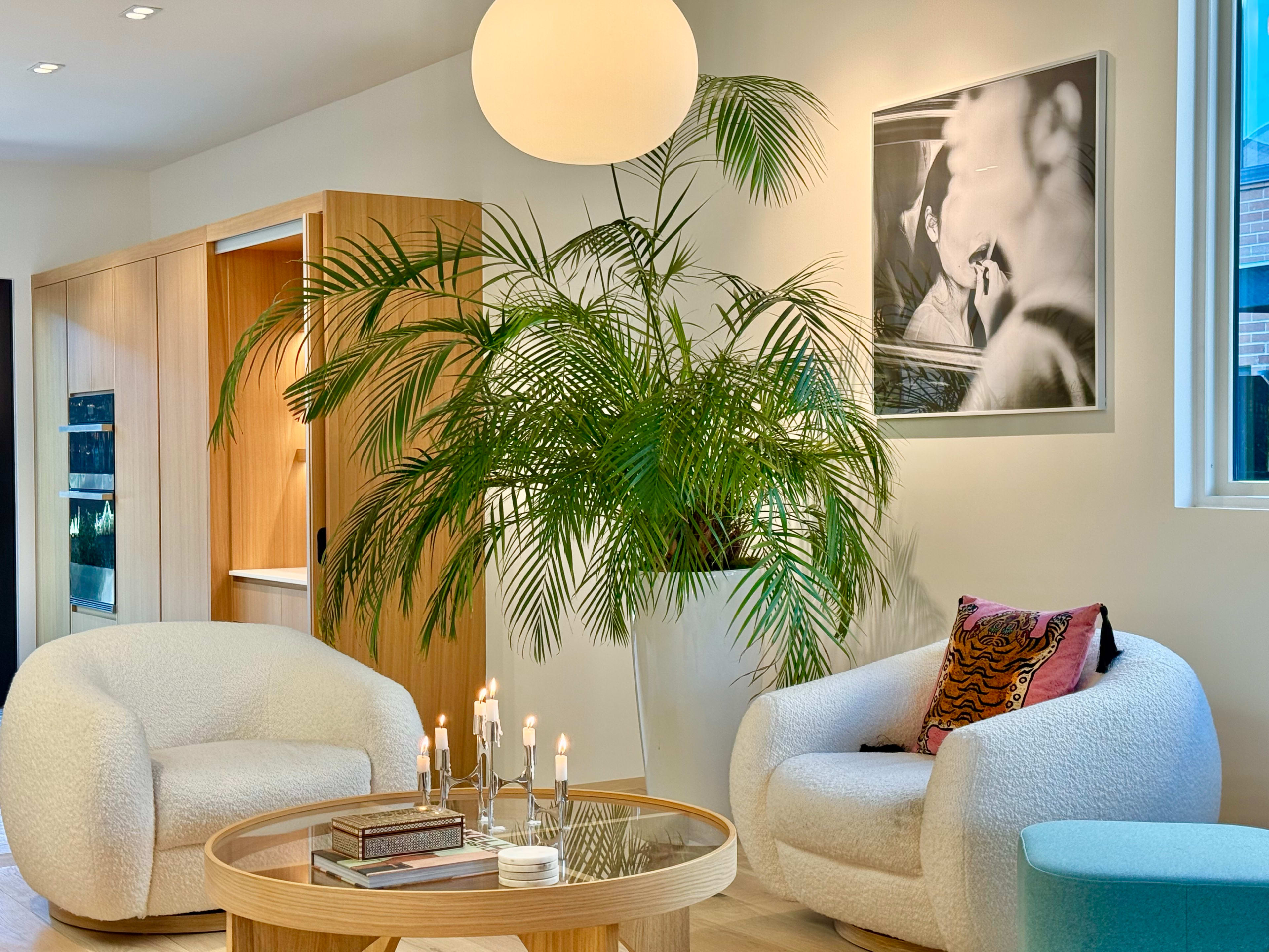 The image shows a modern living room with two cozy chairs, a round table adorned with candles and books, and a large potted plant beside a black-and-white photo on the wall.