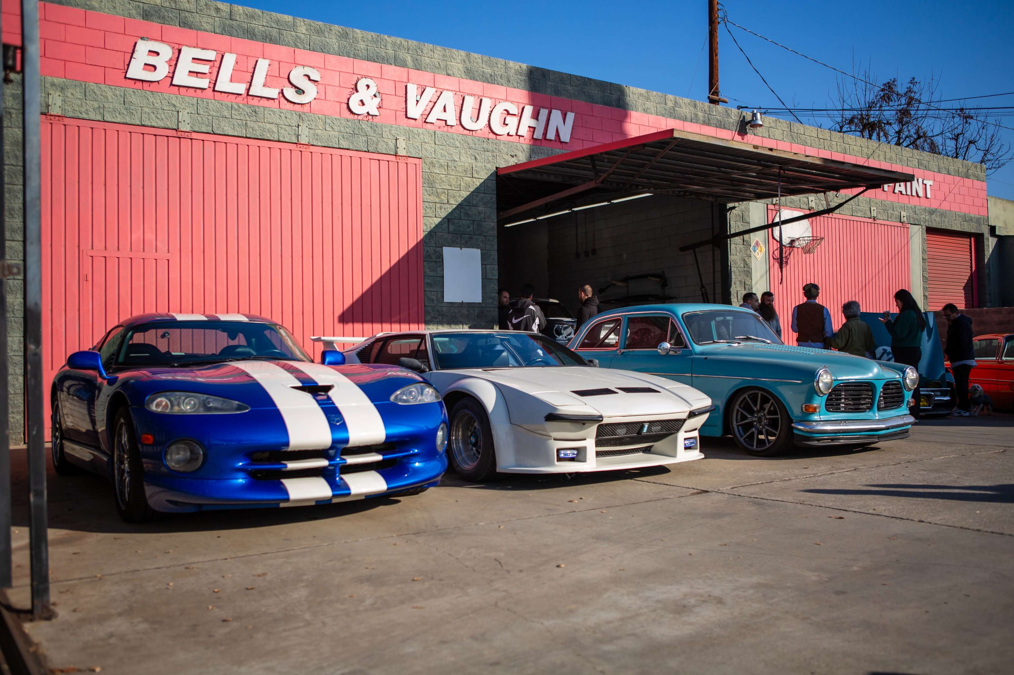 Three cars, including a blue sports car with white stripes, a white modified vehicle, and a vintage blue car, are parked outside a building with a red sign reading "BELLS & VAUGHN."