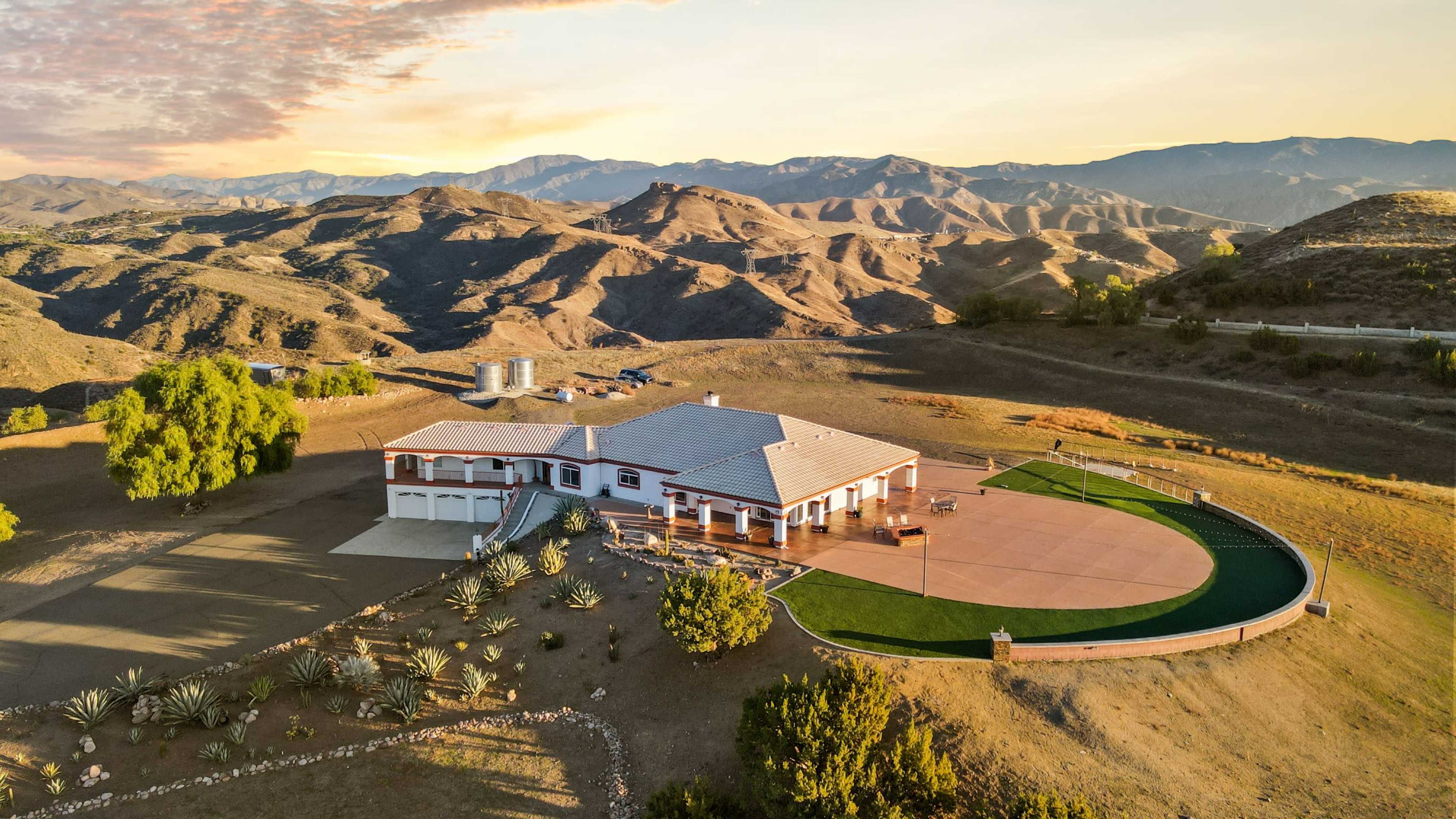 A large, single-story home surrounded by rolling hills and sparse vegetation, with a golf putting green and a parking area visible.