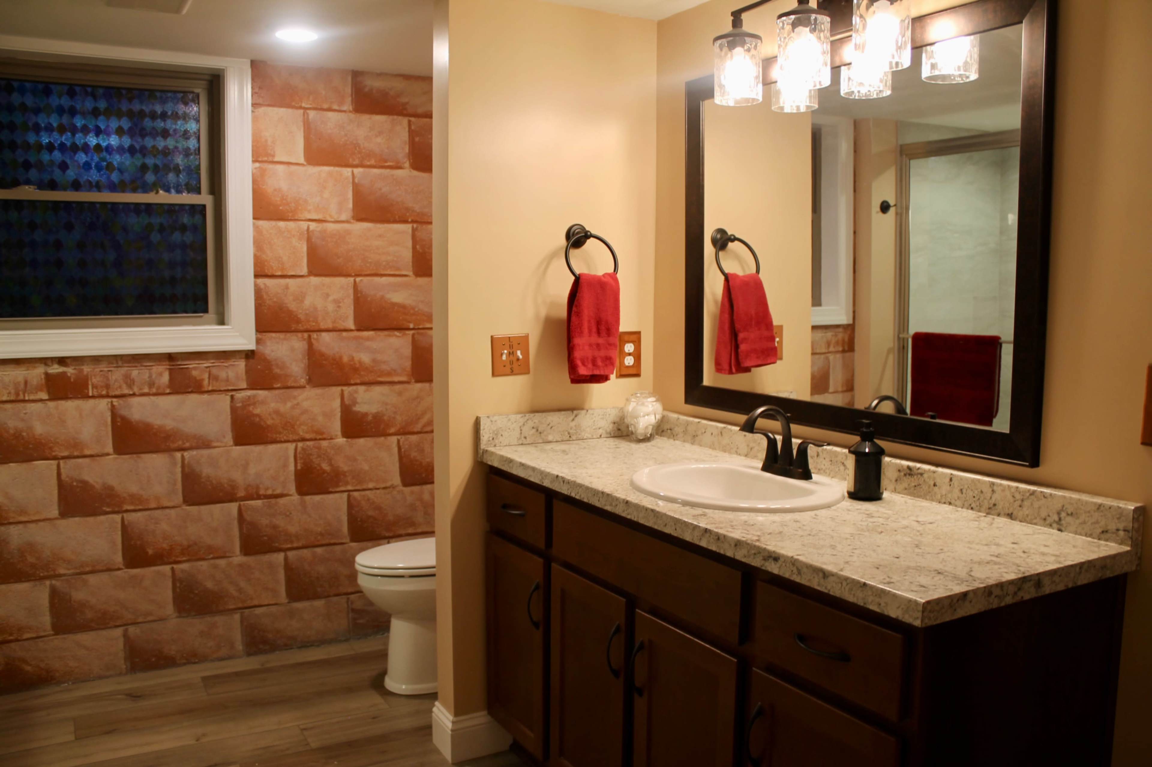 The image shows a bathroom featuring a double vanity with a marble countertop, a large mirror, and a toilet, complemented by a wall with orange brick-like tiles.