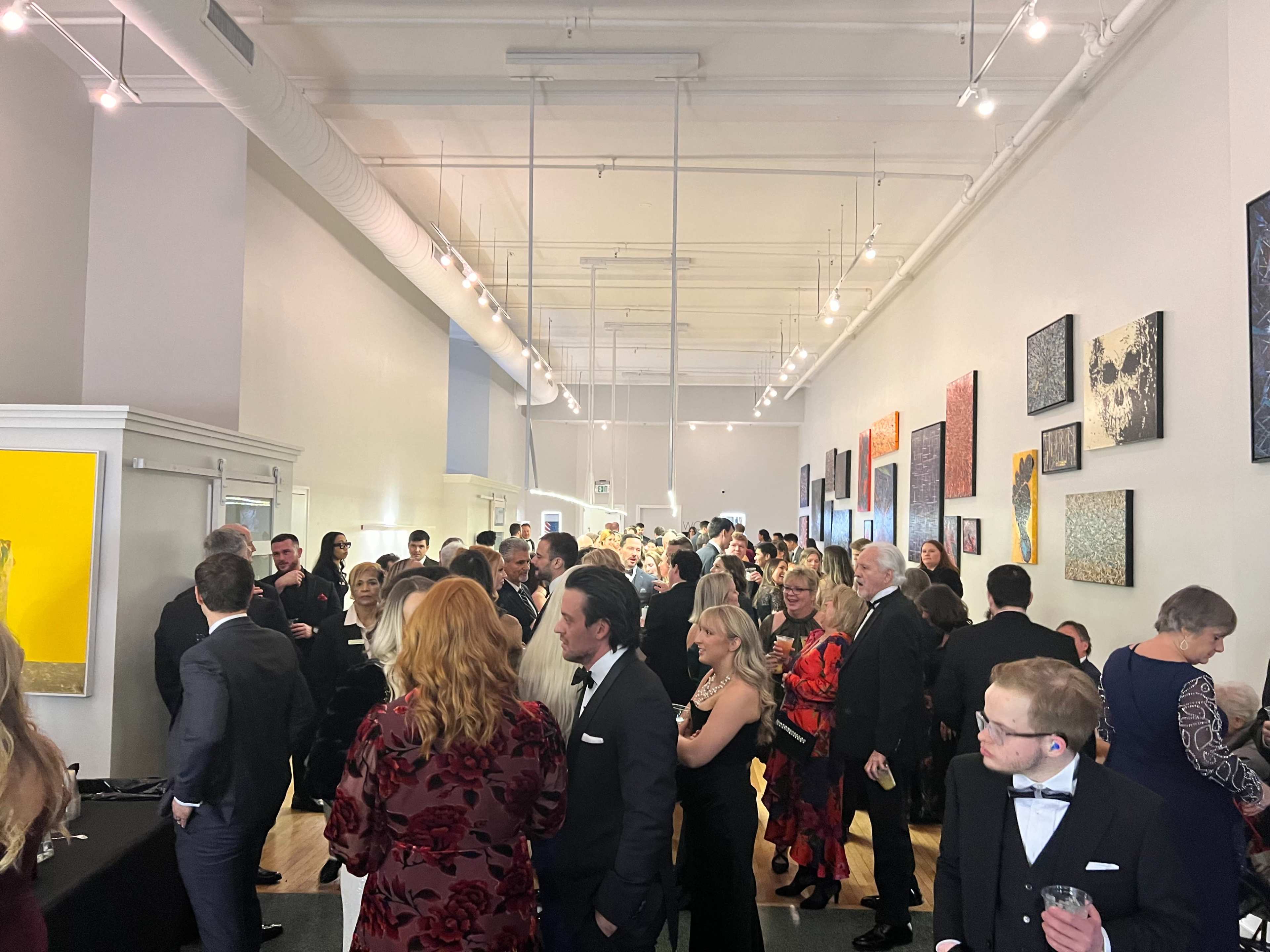 A crowded art gallery during an event, with guests mingling and viewing artworks displayed on the walls.