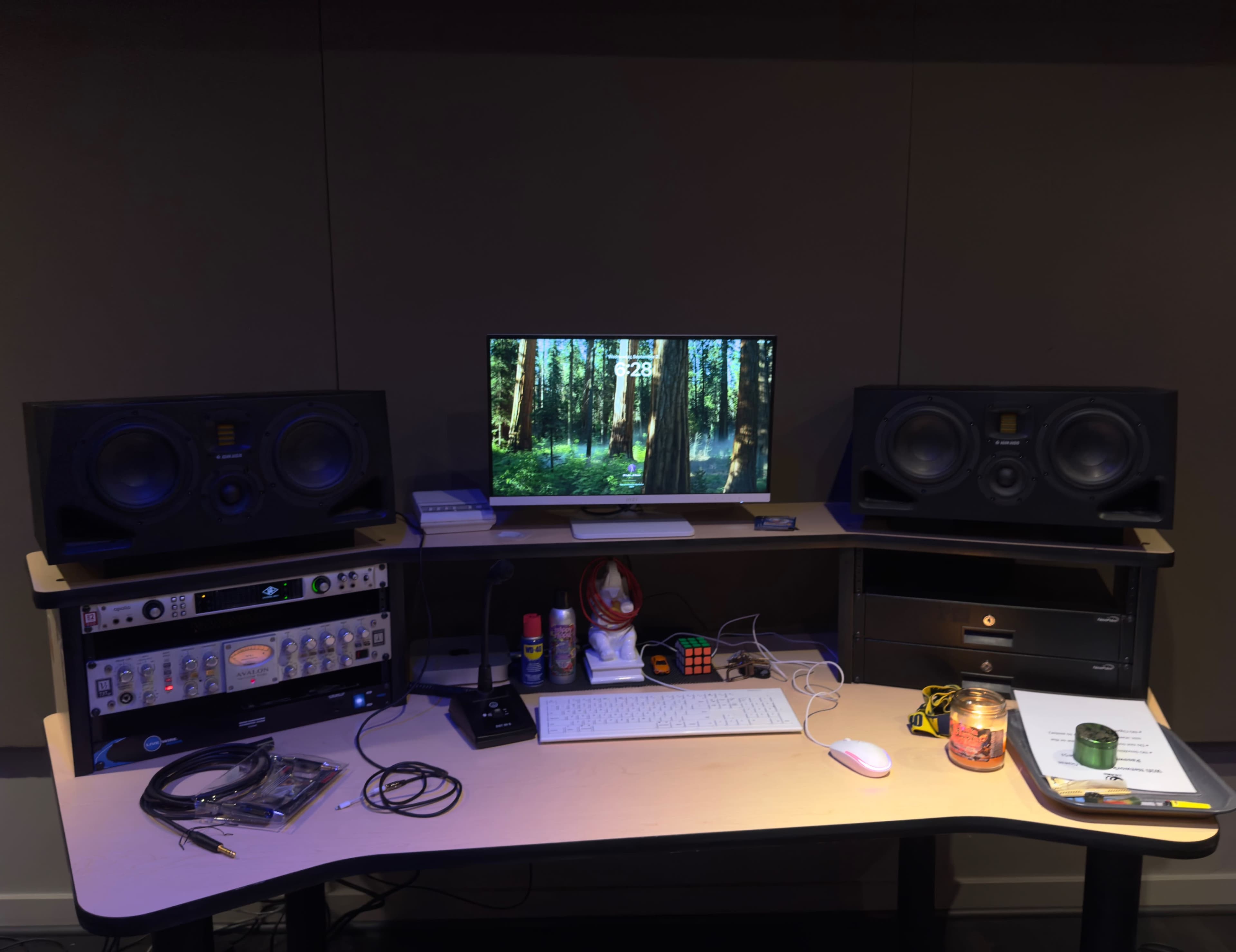 A recording studio desk features two large speakers, a computer monitor displaying a forest scene, and various audio equipment alongside cables and a lit candle.