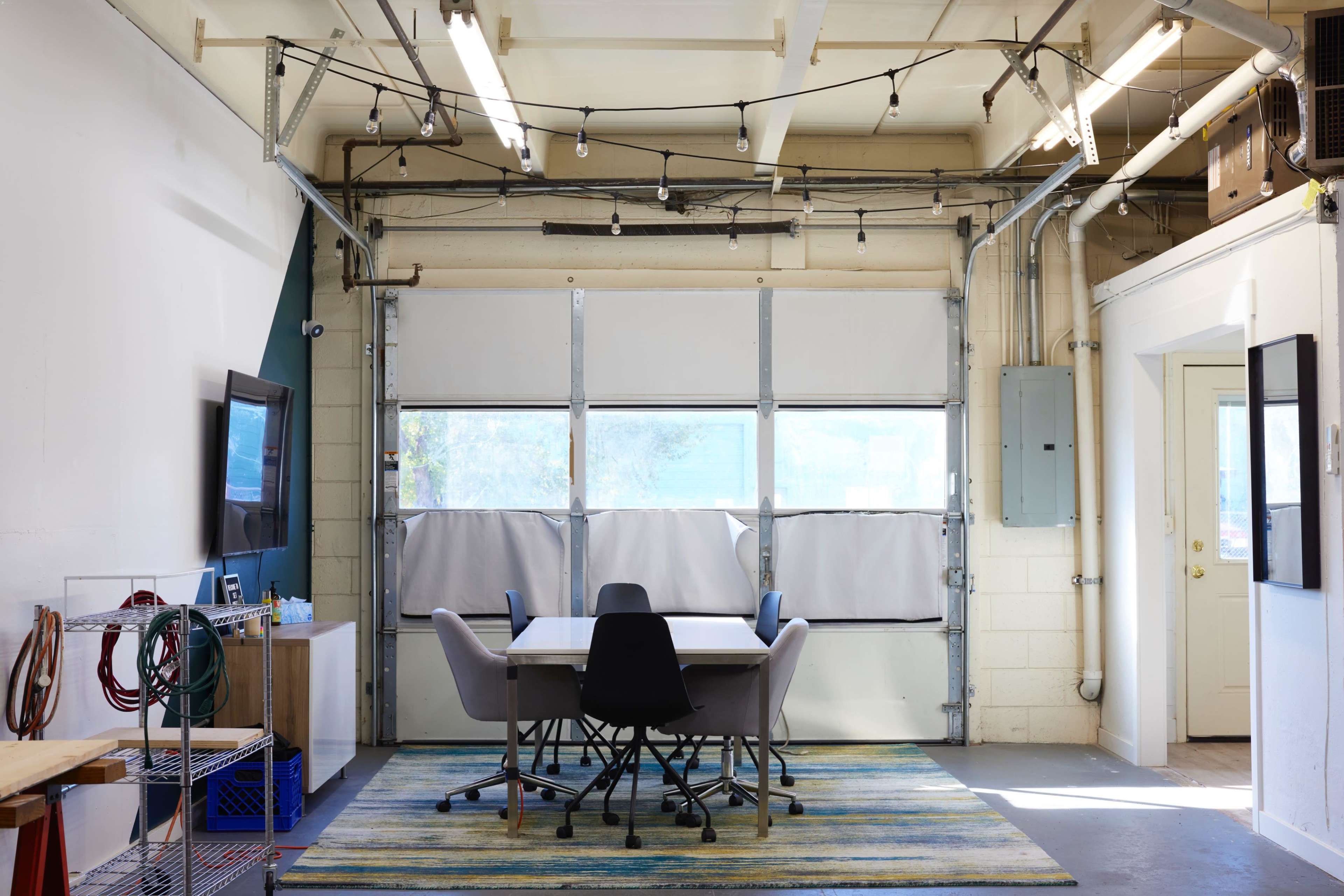 A workspace with a large table surrounded by chairs, located in a garage-like setting with large windows and exposed electrical features.