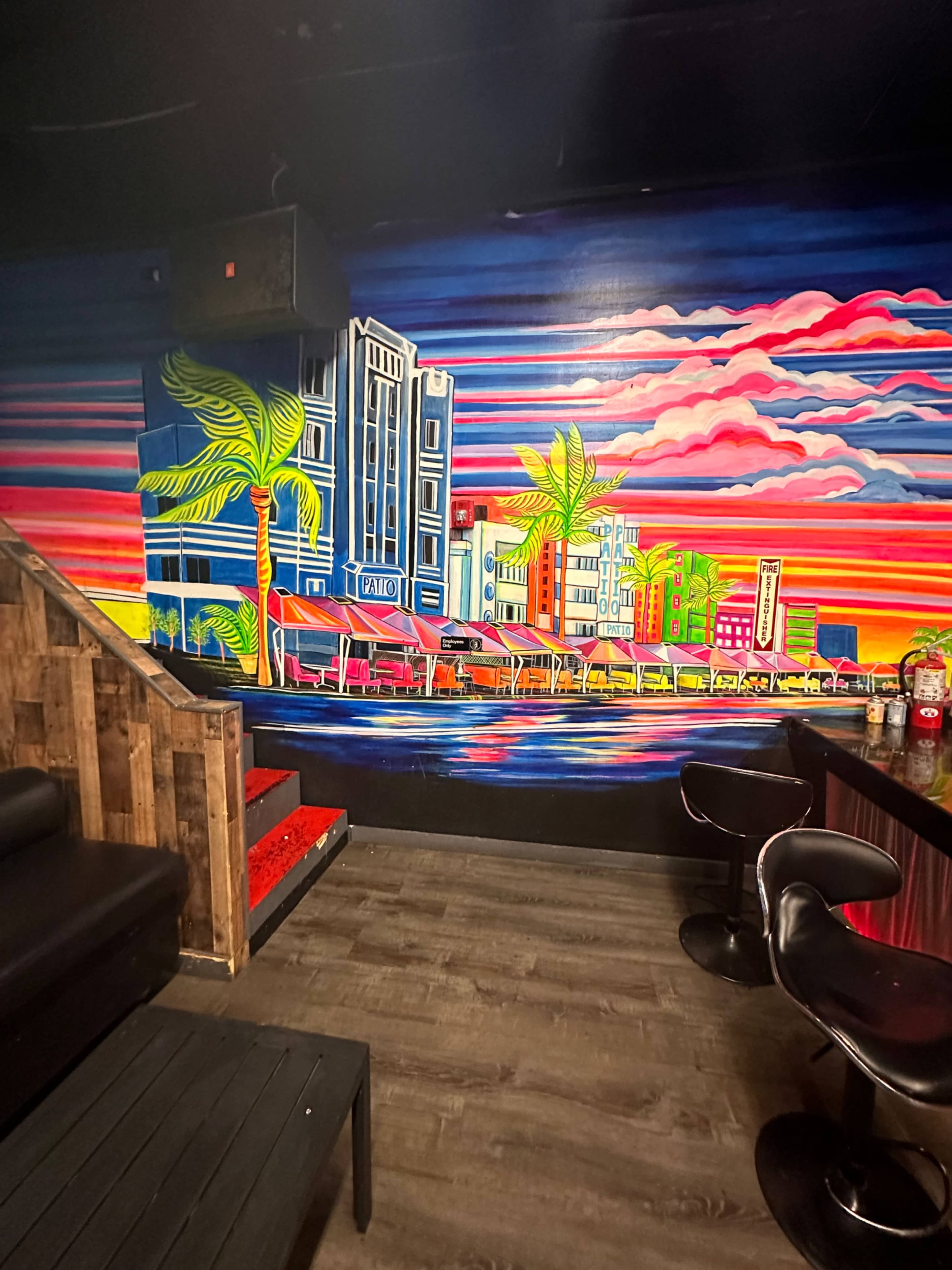A vibrant mural featuring a colorful cityscape with palm trees, set against a striped sunset backdrop, inside a bar area with dark furniture.