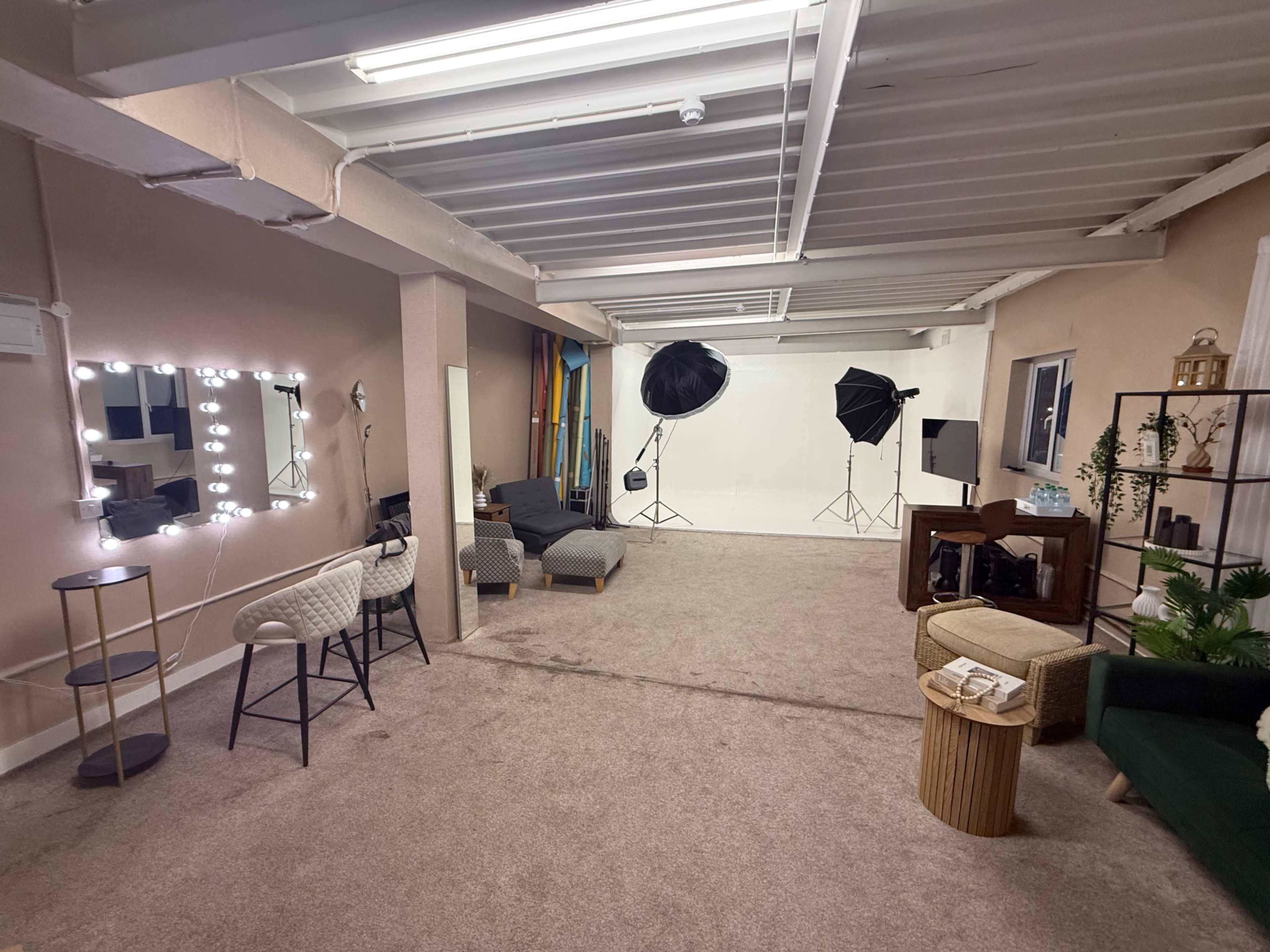 The image shows a spacious room with a carpeted floor, featuring a seating area with chairs and a small table, and equipment set up for photography, including light stands and backdrops.