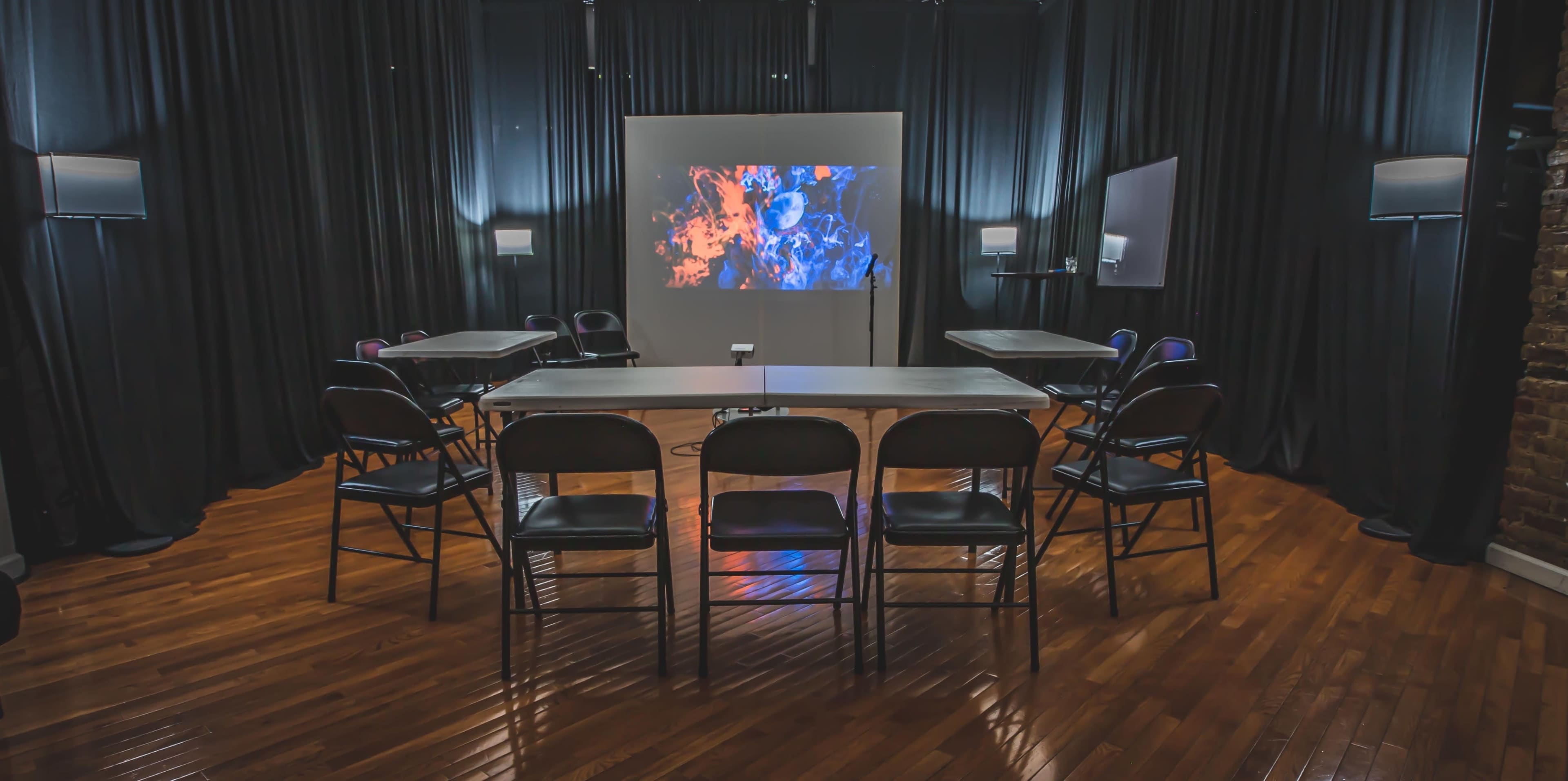 A modern meeting room features a central table surrounded by chairs, with a large screen displaying colorful visuals against a backdrop of black curtains.