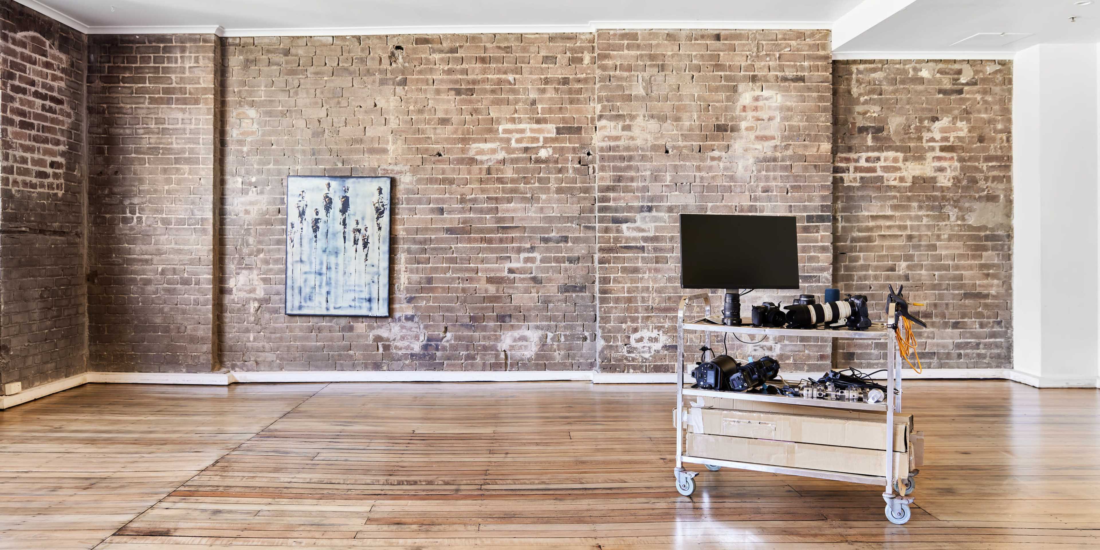 A large, empty room features exposed brick walls, a wooden floor, a piece of artwork, and a media cart with a monitor and camera equipment.