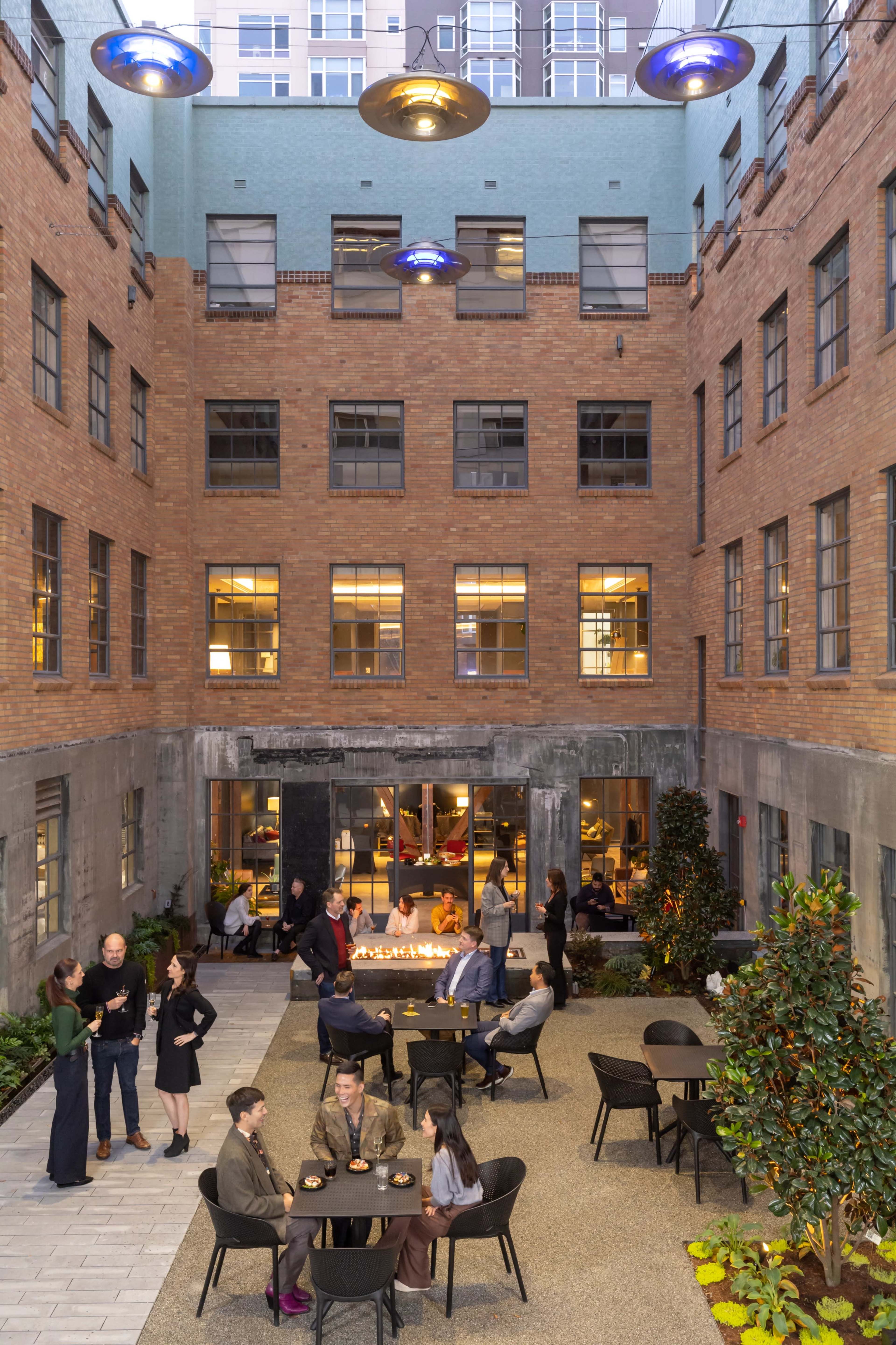 A group of people socializes around tables in a courtyard surrounded by a brick building, with a fire feature and modern lighting.