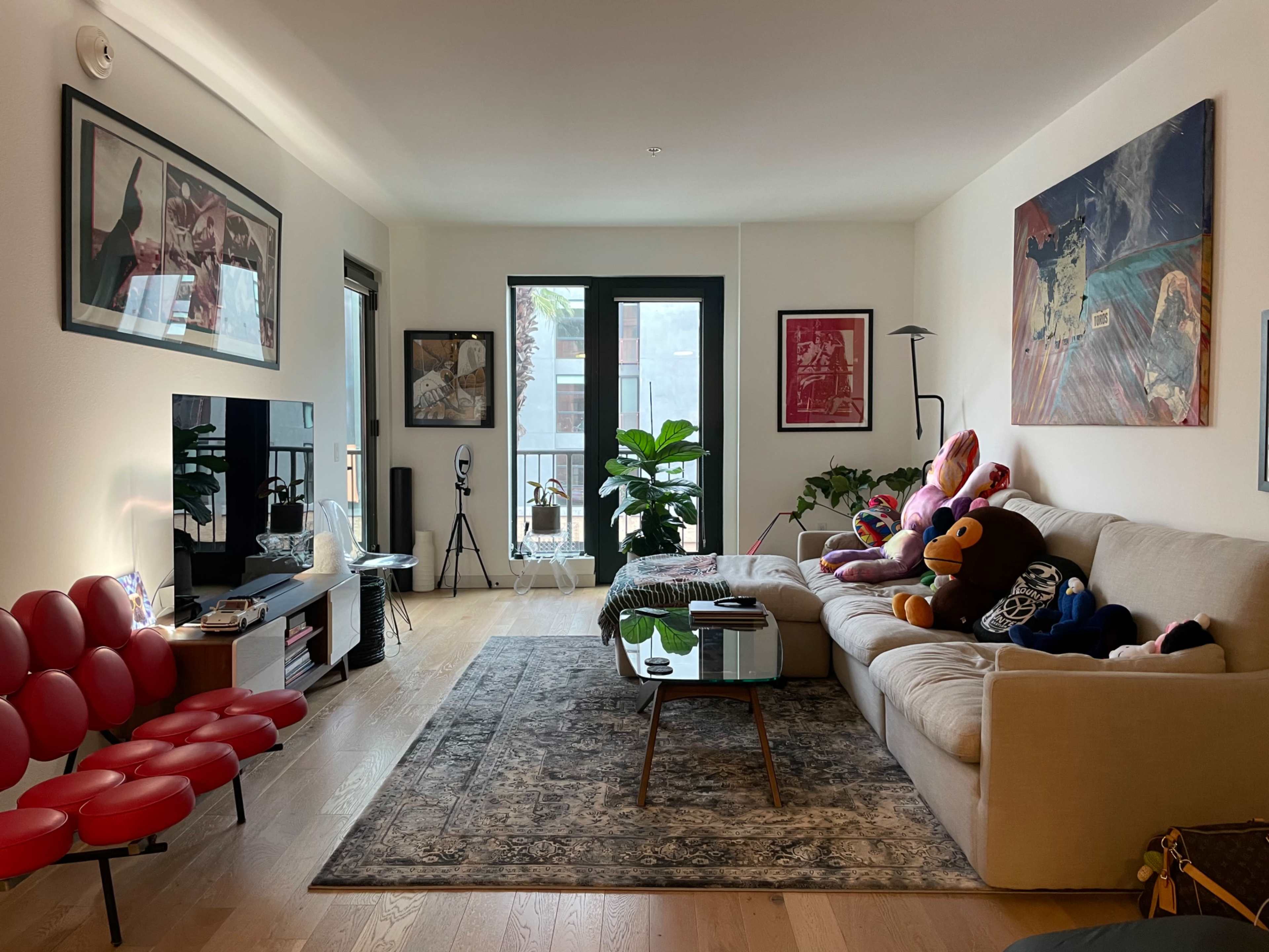 The image shows a modern living room with a beige sofa, a decorative rug, various plush toys on the couch, and large windows allowing natural light to enter.