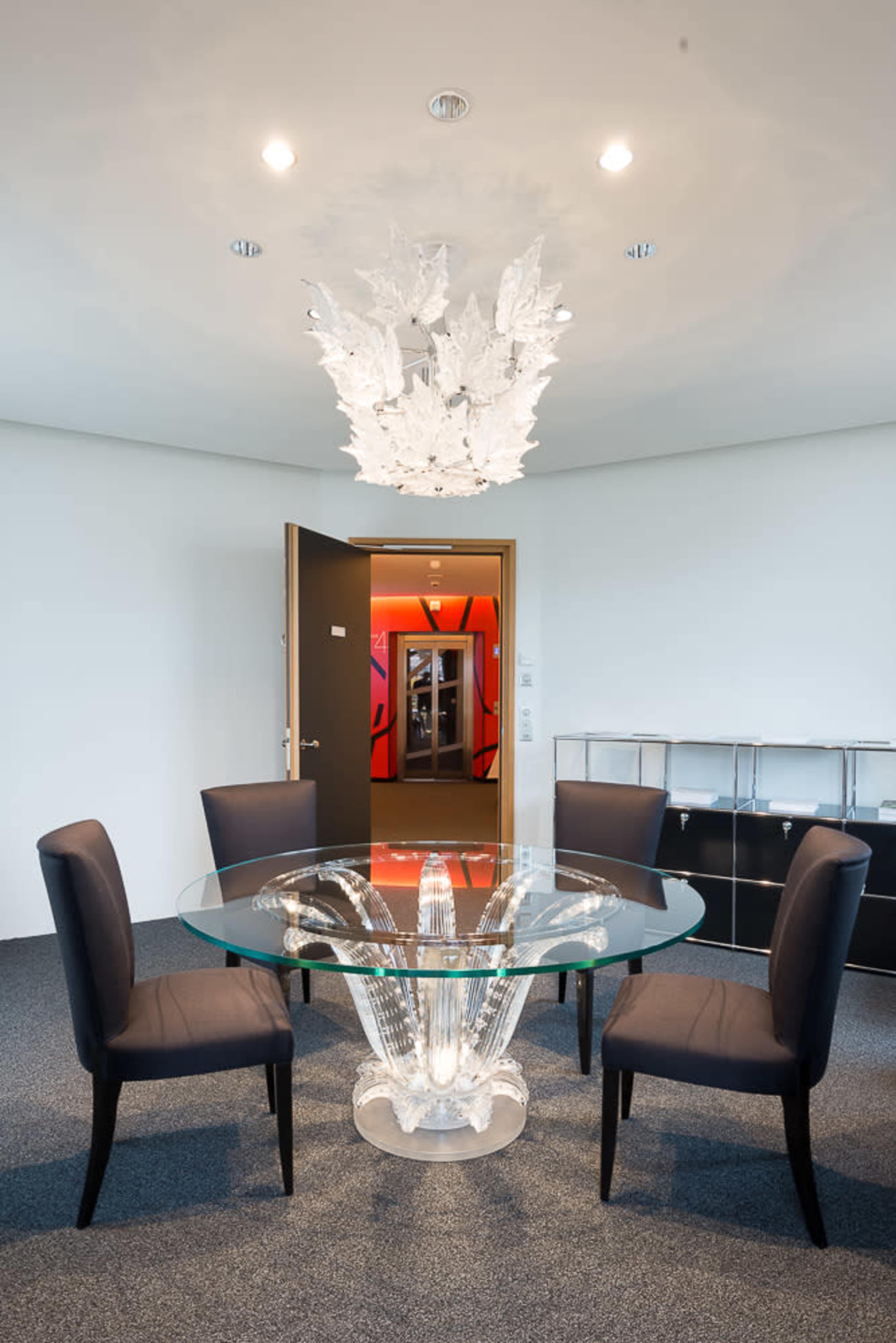 A modern dining area featuring a glass table with an intricately designed base and four black upholstered chairs, illuminated by a decorative ceiling light.