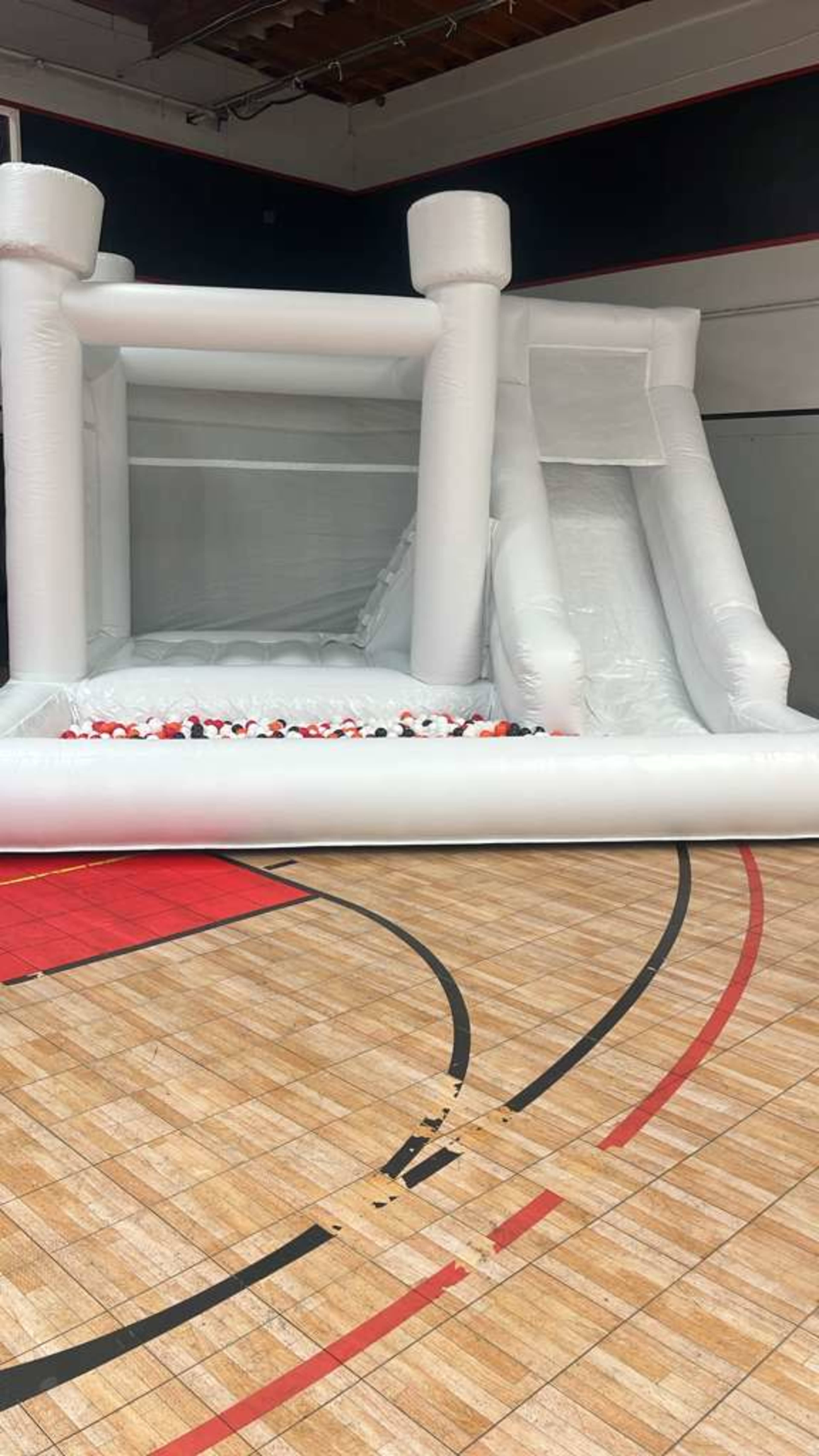 The image shows a large inflatable bounce house featuring a slide and a ball pit, set up on a hardwood gym floor.