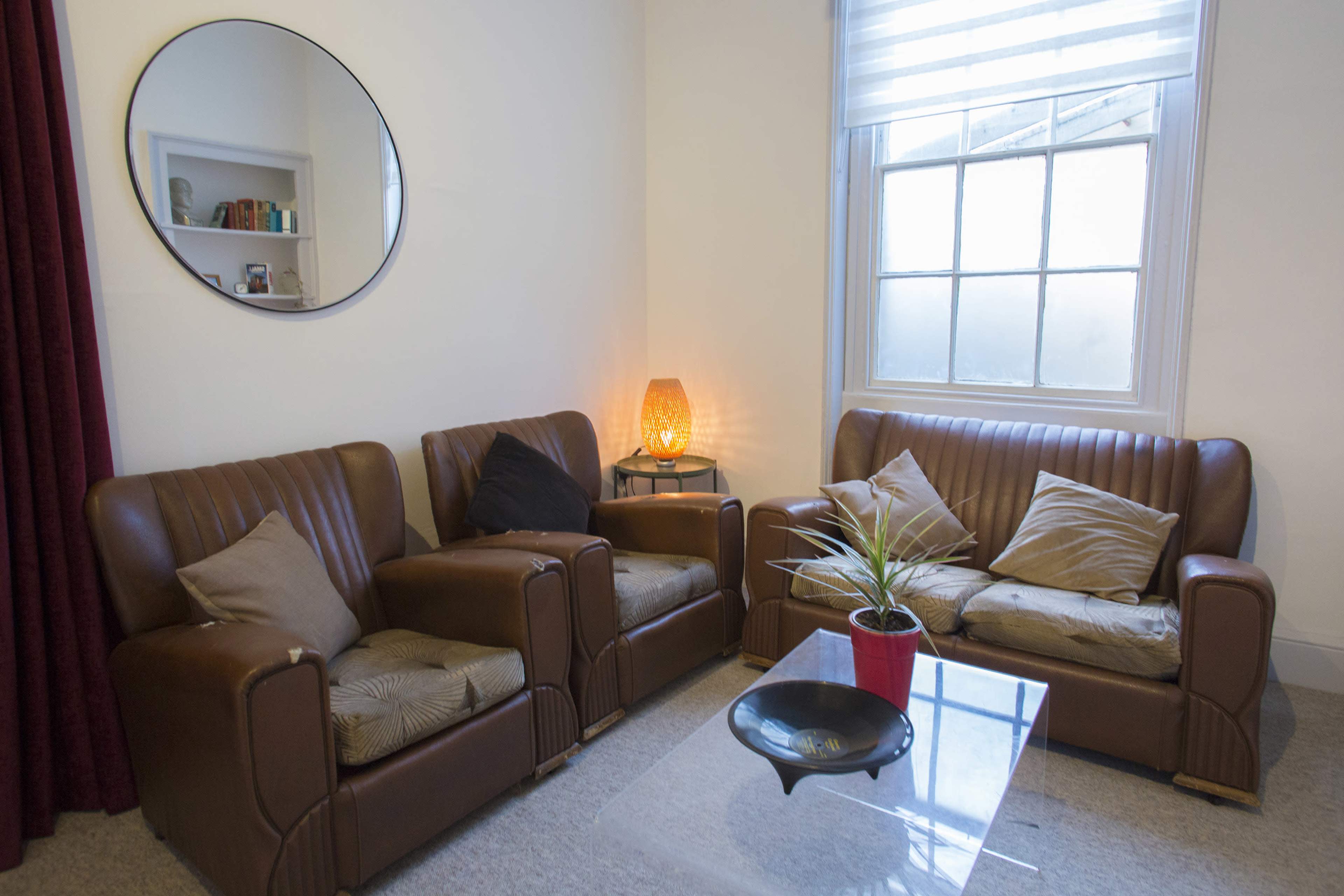 A living room corner with two brown leather sofas, a glass coffee table, a potted plant, and a round mirror reflecting a window.