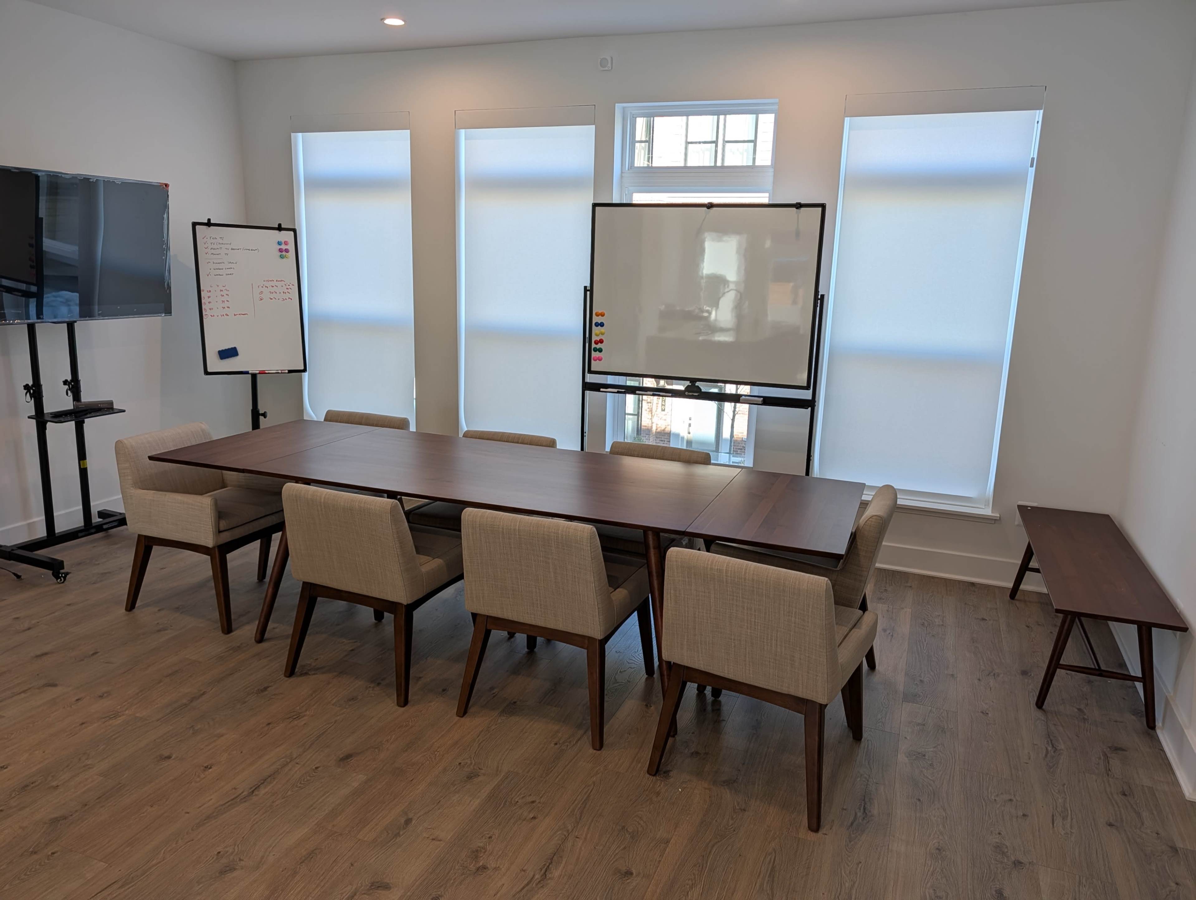 A long, rectangular table surrounded by eight upholstered chairs is positioned in a well-lit meeting room with large windows and a whiteboard.