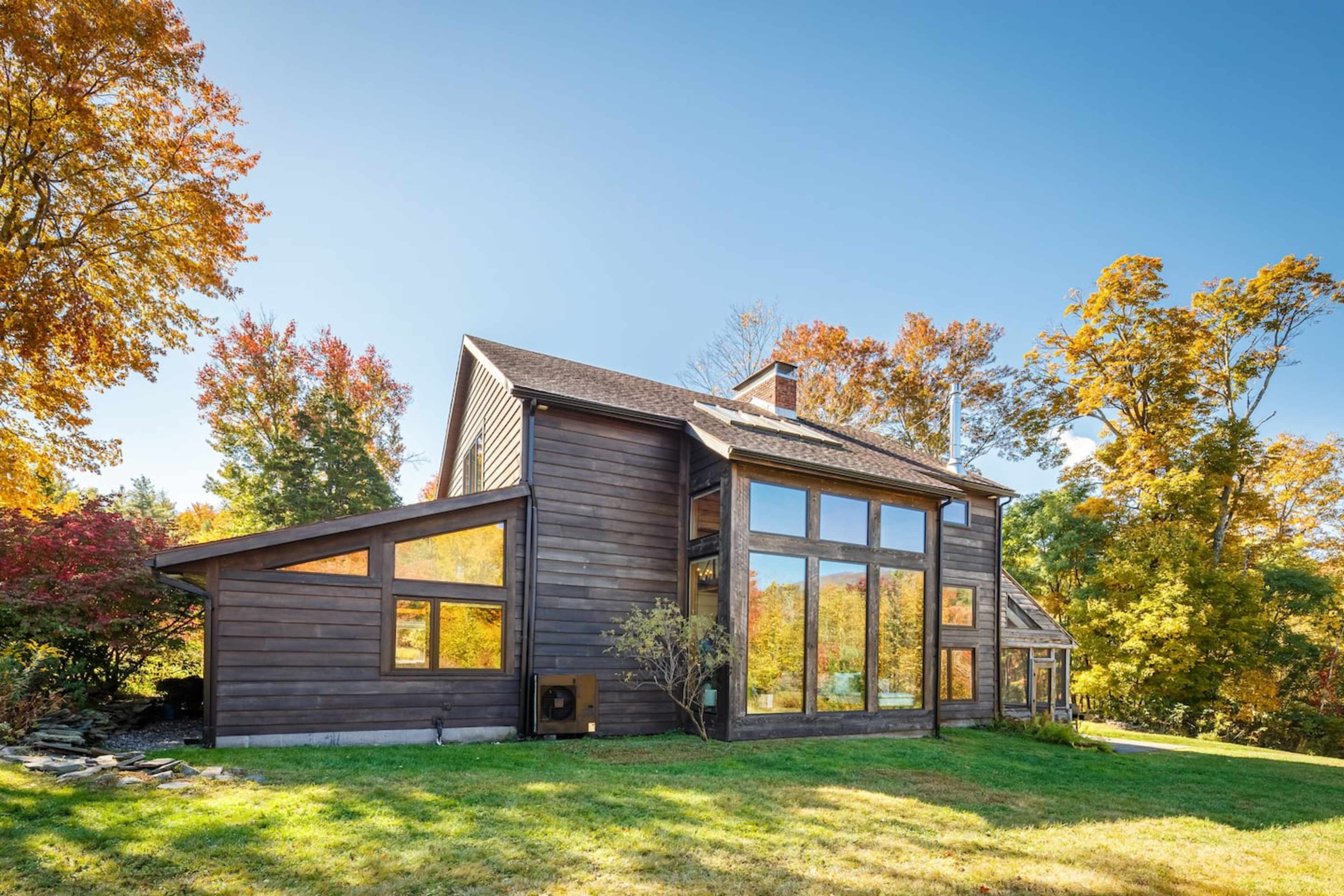 A modern wooden house with large windows is surrounded by colorful autumn trees and a clear blue sky.