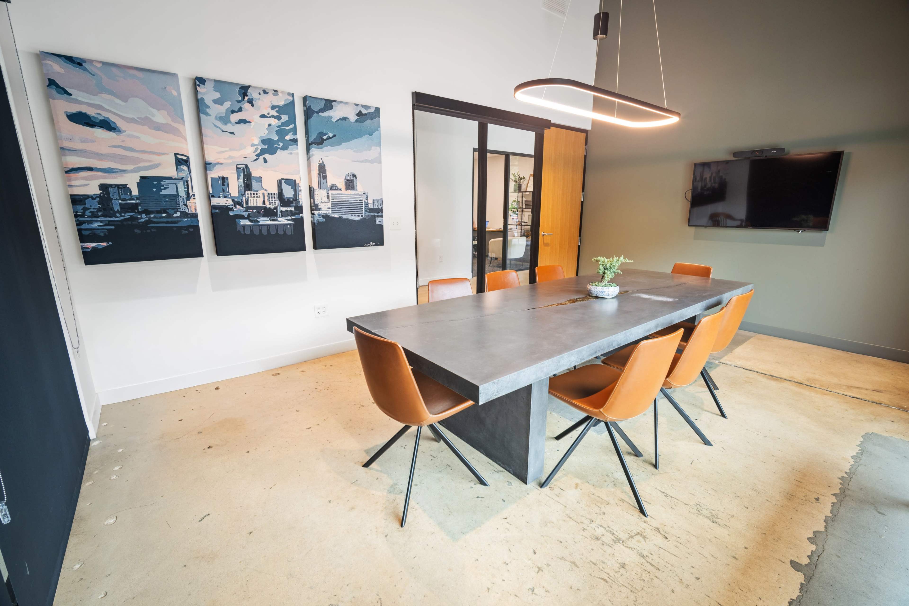 A modern conference room features a large gray table surrounded by orange chairs and three abstract cityscape paintings on the wall.
