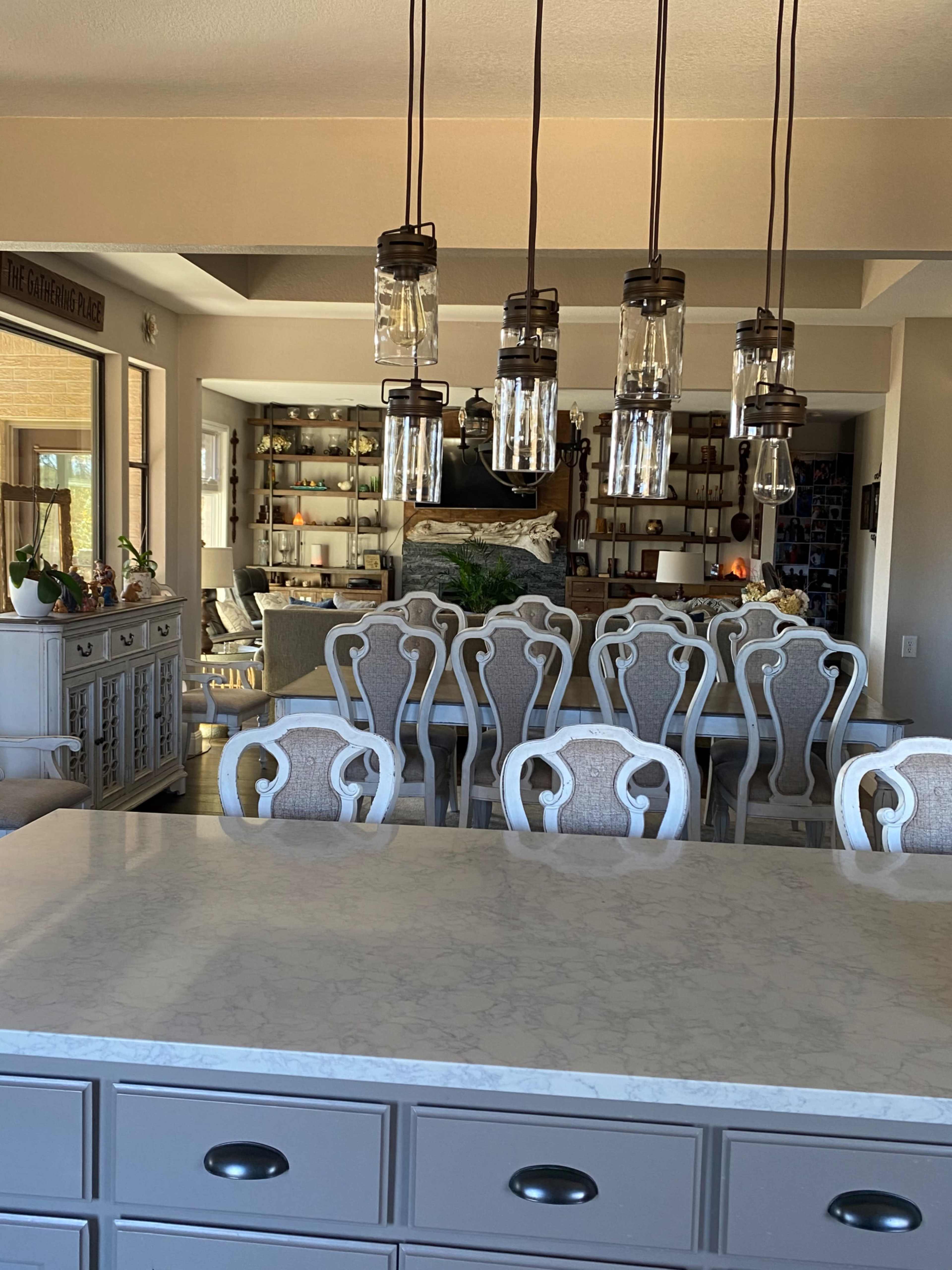A dining area with a long table and upholstered chairs is illuminated by pendant lights above, while a decorative sideboard and shelves are visible in the background.