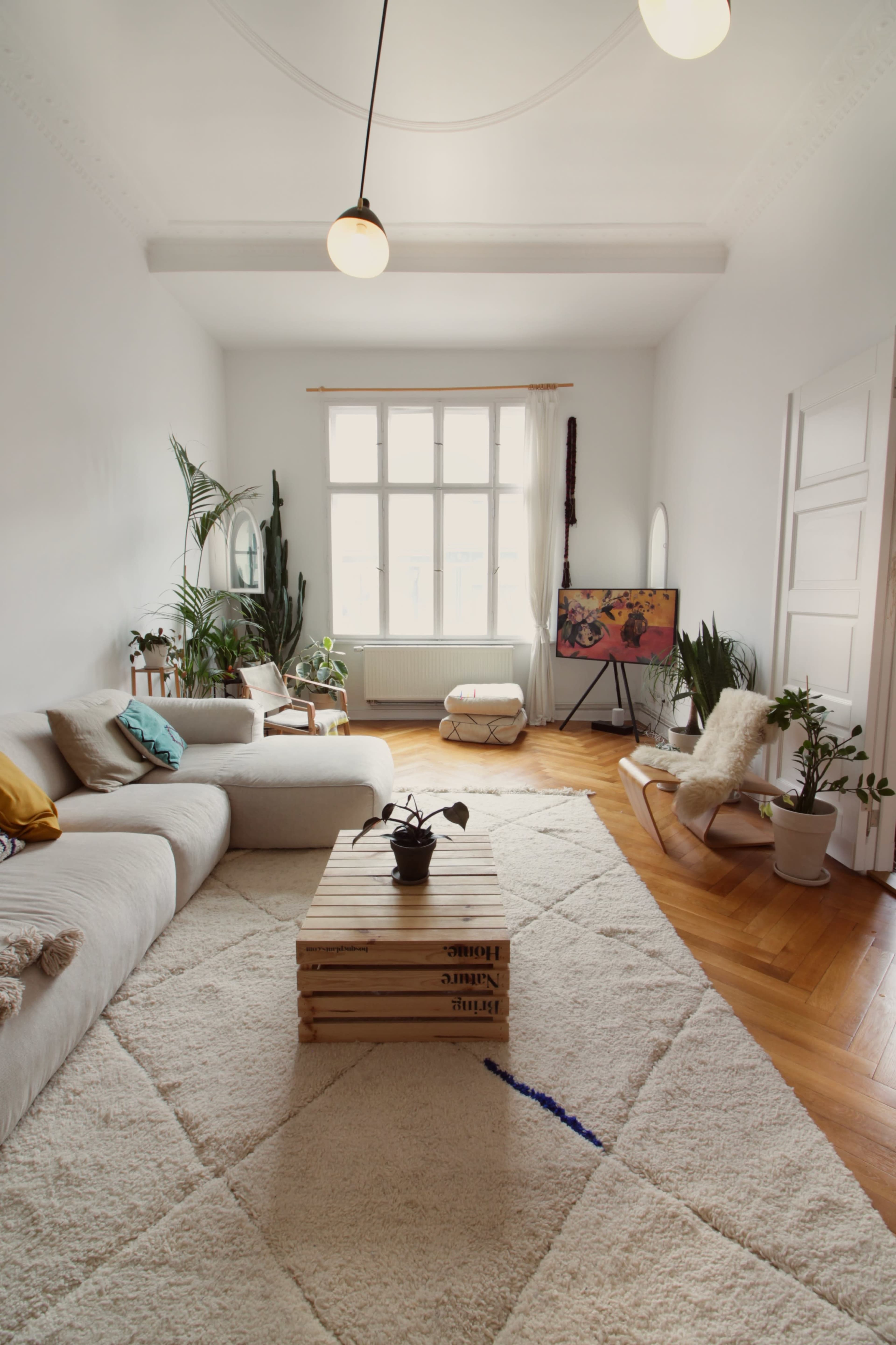 The room features a light, spacious living area with a large sectional sofa, wooden coffee table, plants, and a wall-mounted artwork.