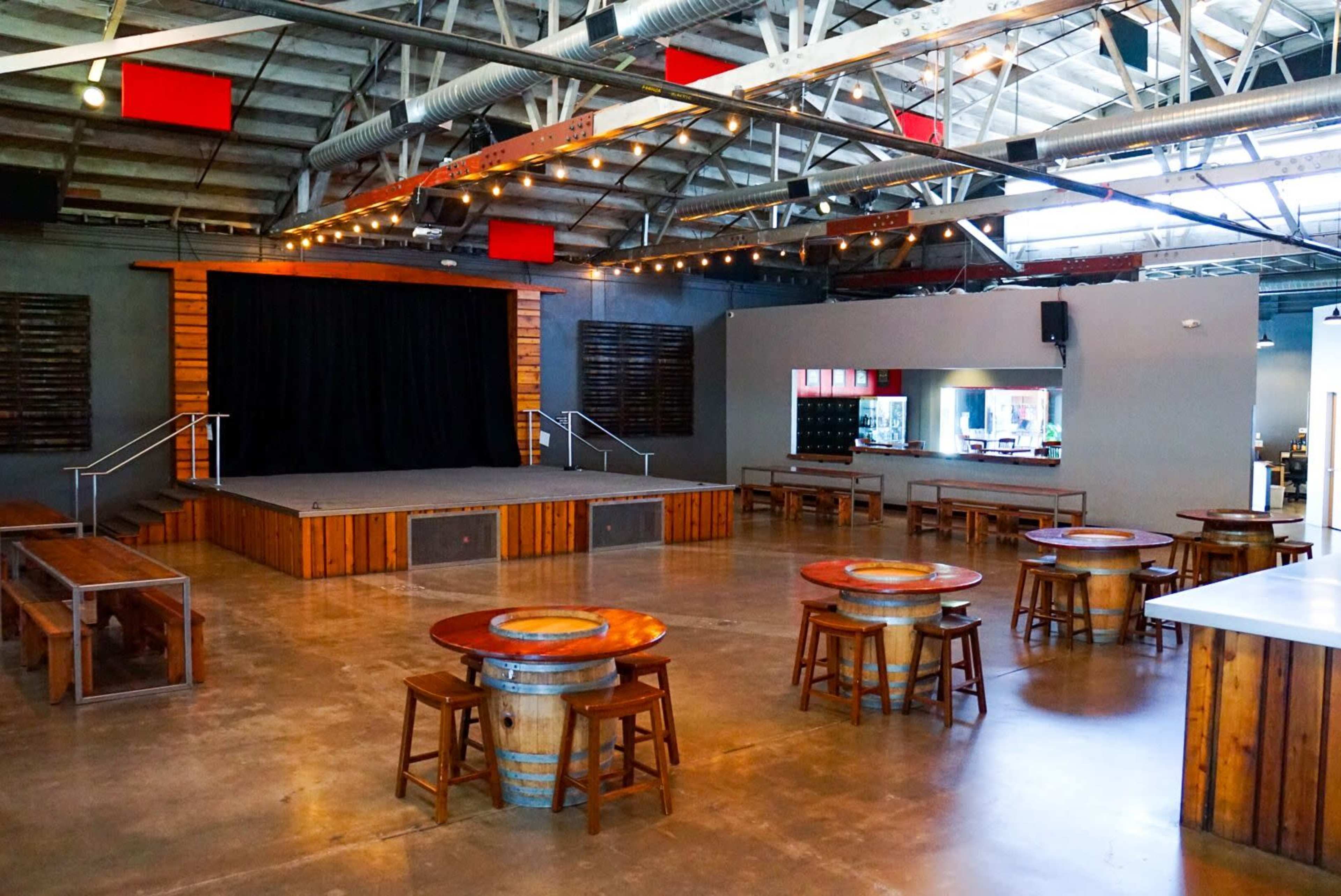 The image shows a spacious event venue with a wooden stage at one end, surrounded by round tables made from barrels and wooden stools.