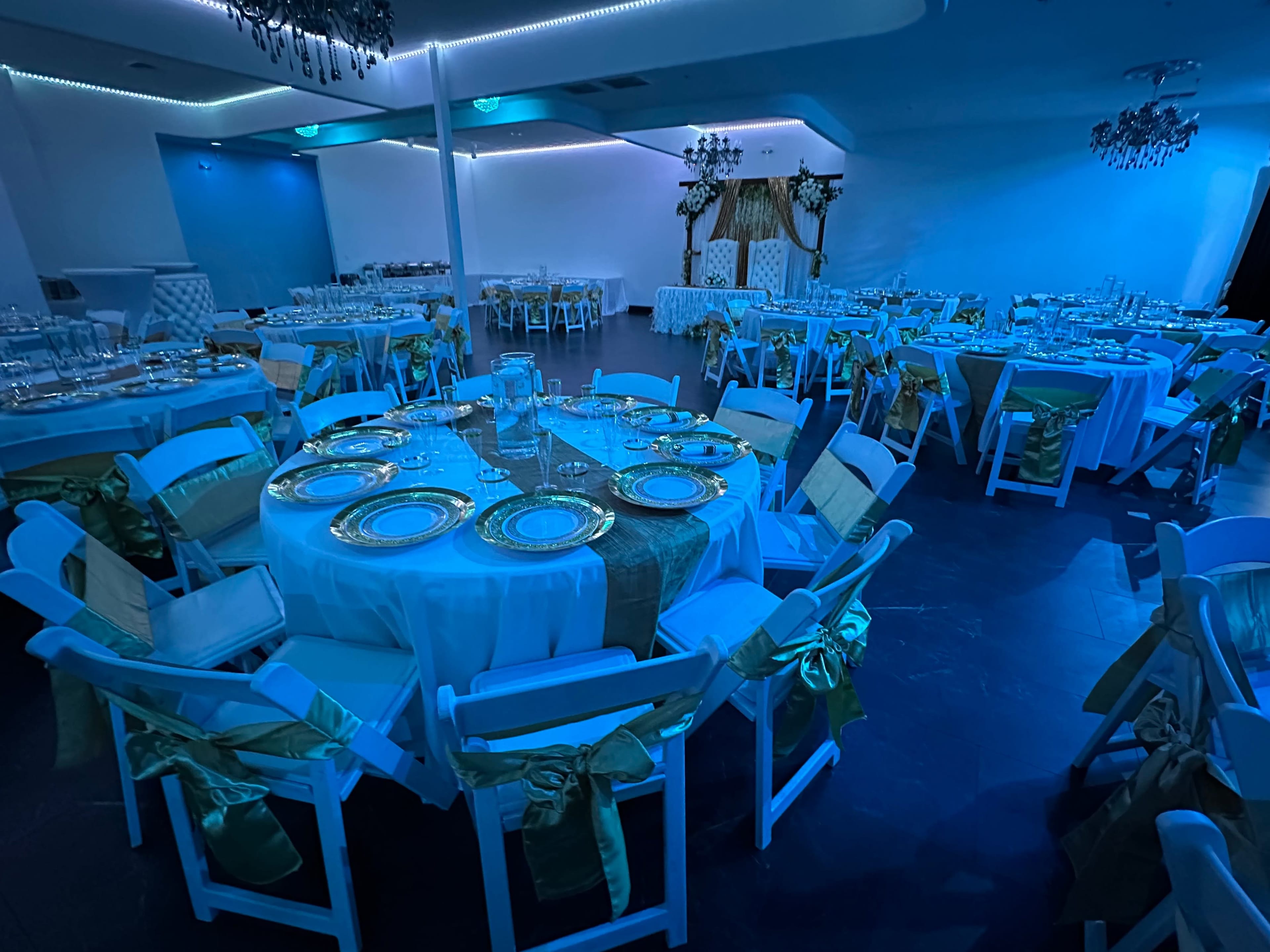 The image shows a banquet hall set up for an event, featuring round tables with white tablecloths, gold plates, and green sashes on the chairs, all illuminated by blue lighting.