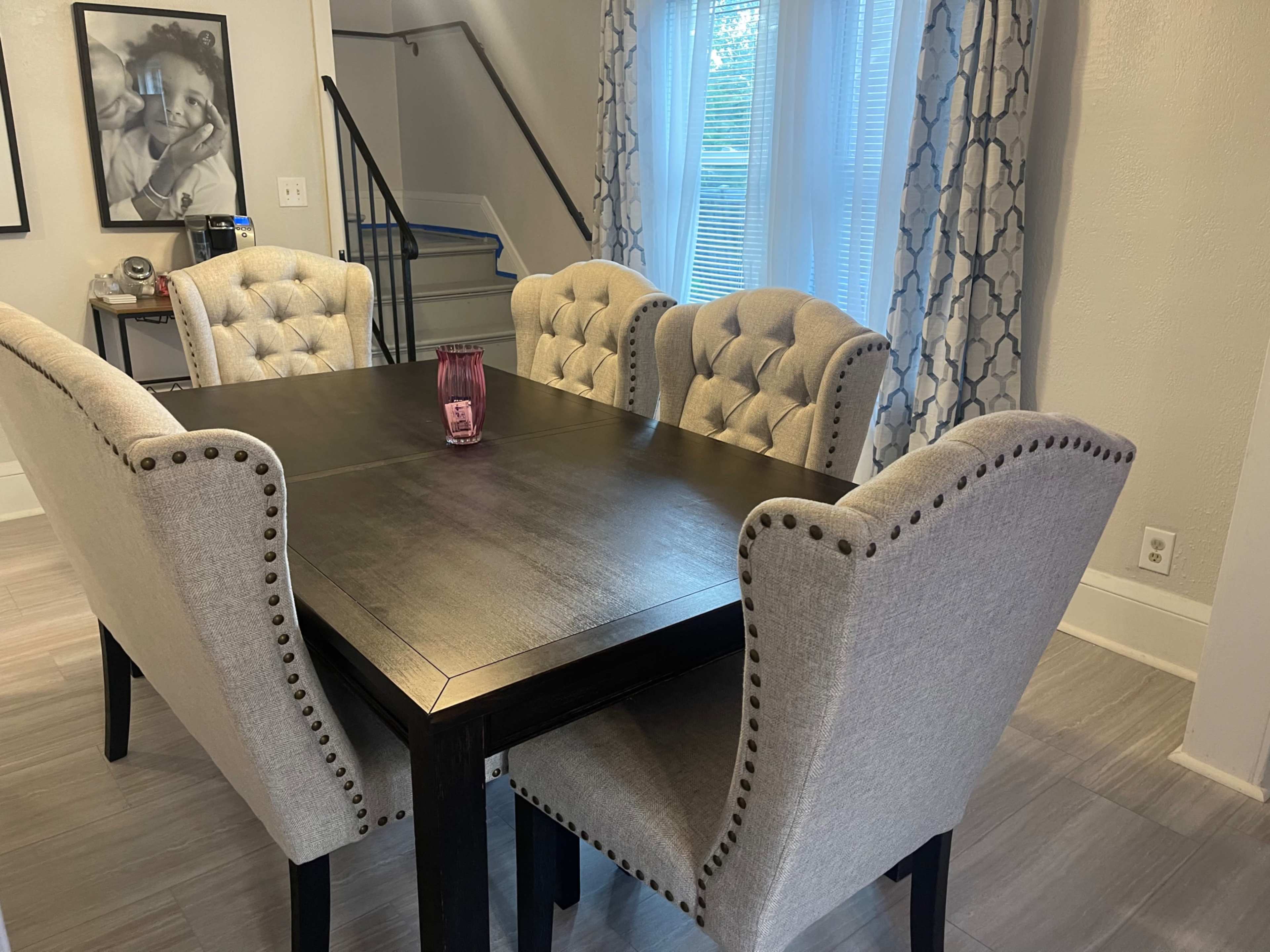 A dining table surrounded by four upholstered chairs sits in a well-lit room with a staircase and window in the background.