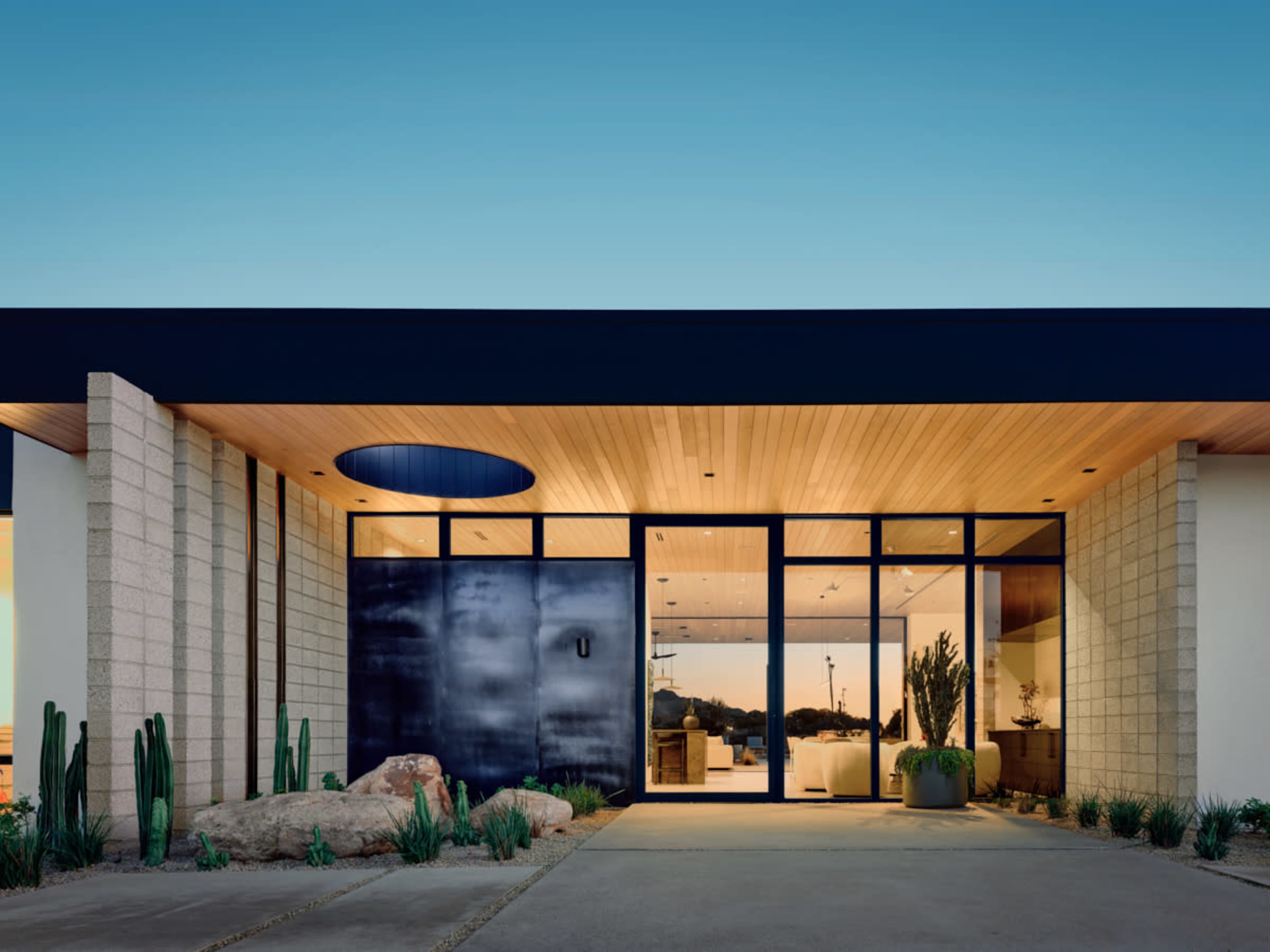 The image shows a modern home with a flat roof, large glass front doors, and natural stone landscaping in the entrance area.
