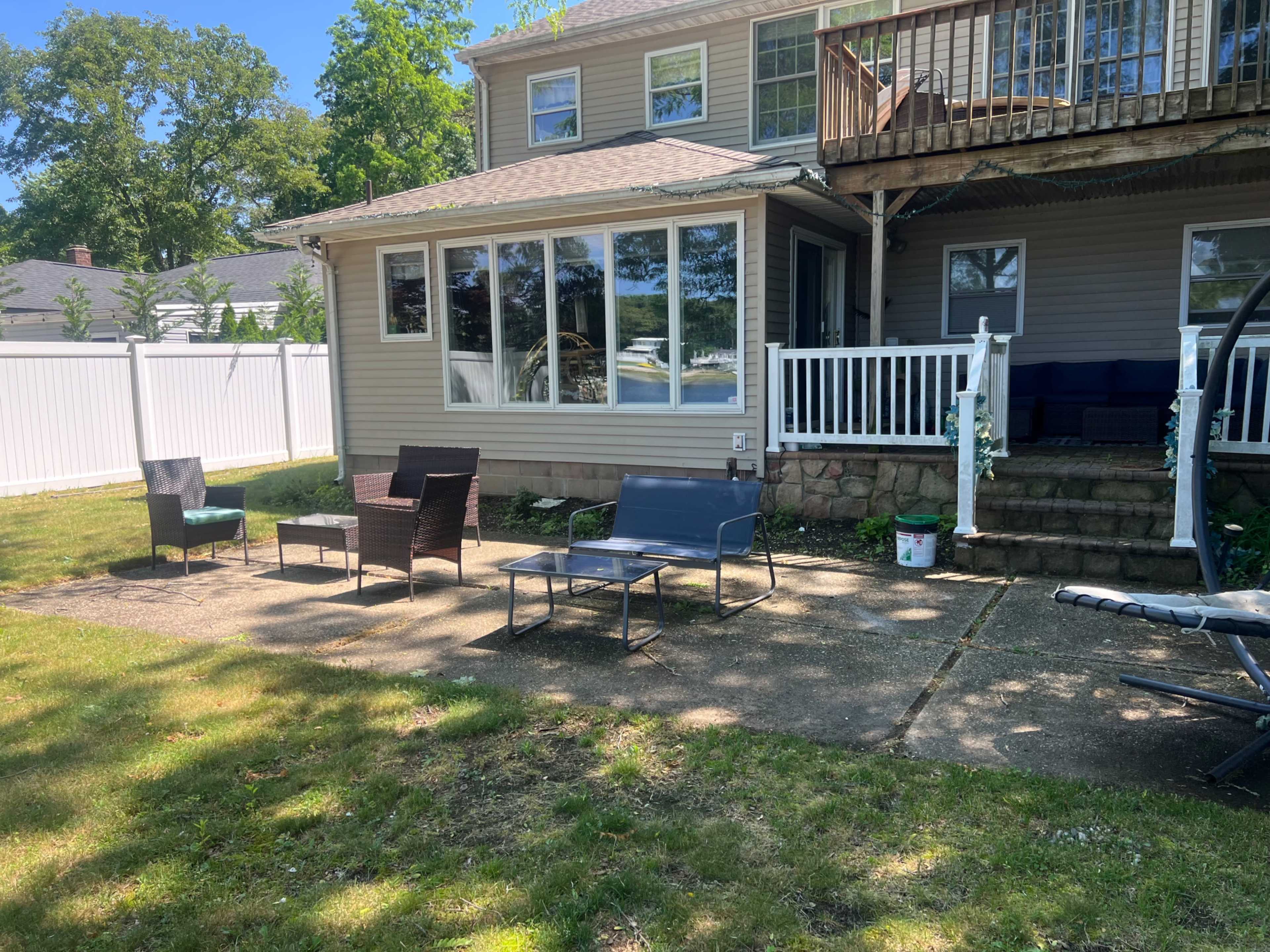 A backyard patio features several chairs and a table, bordered by a white fence and a multi-level house.