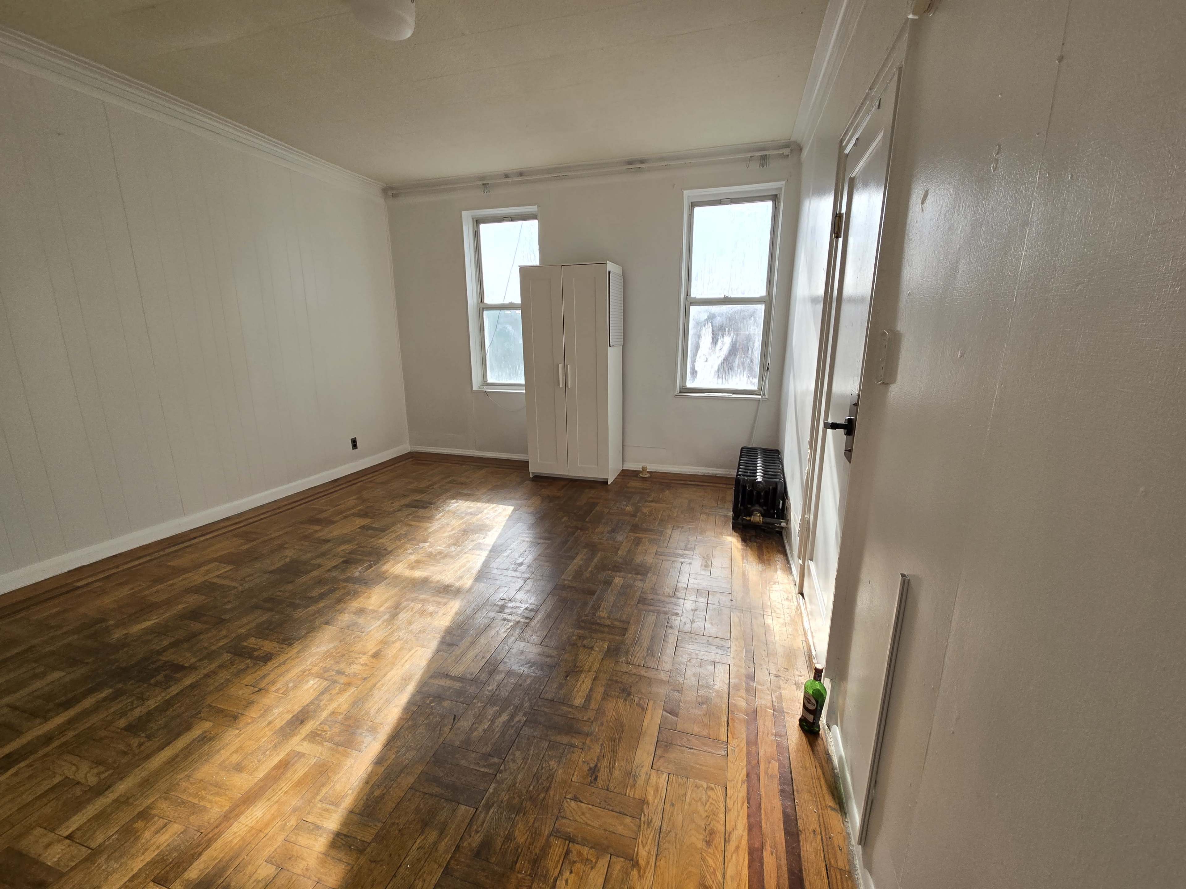 The image shows an empty room with two windows, wooden parquet flooring, and a white cabinet against one wall.