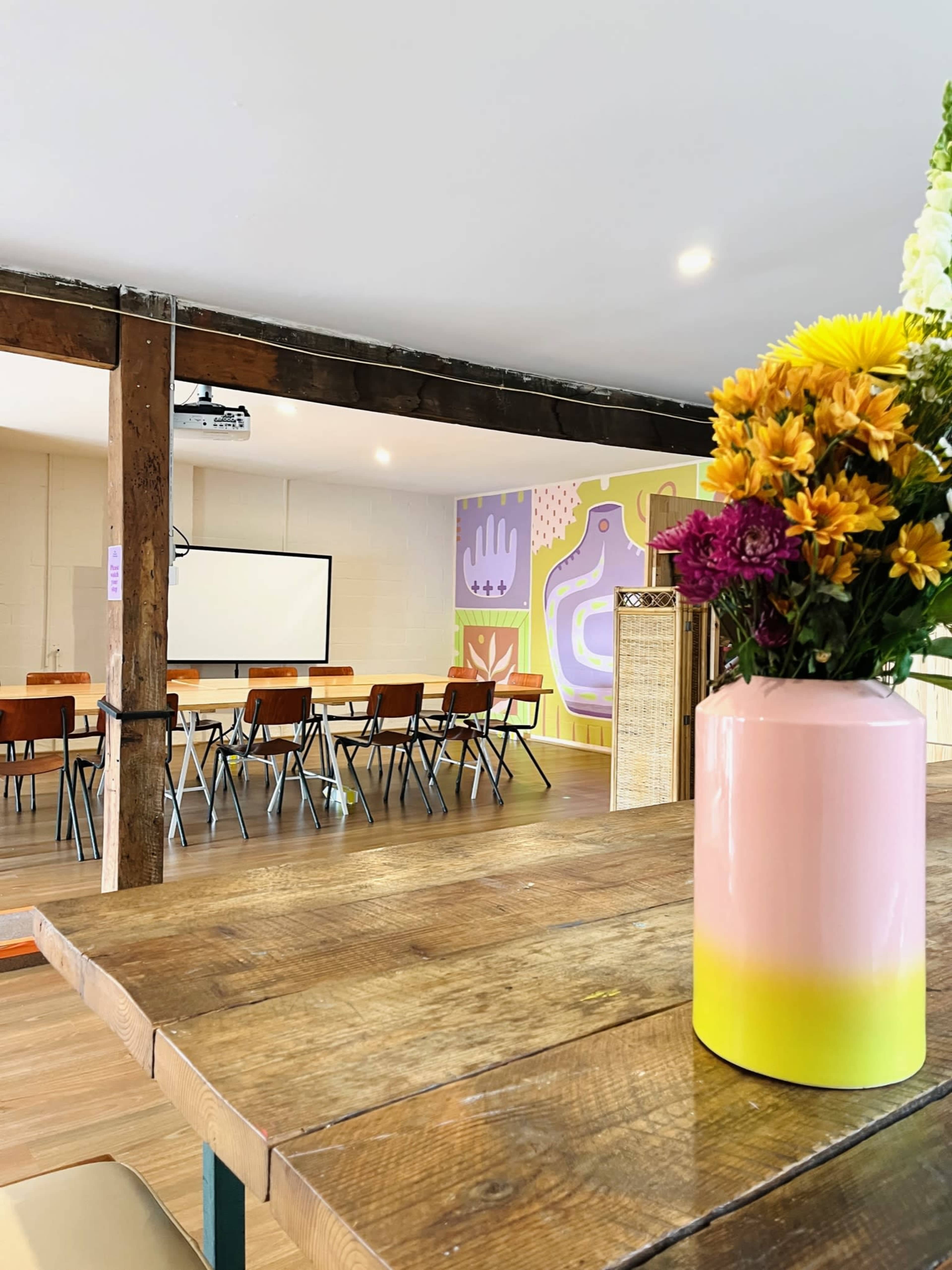 A brightly lit room features a wooden table with a vase of flowers in the foreground, and chairs arranged around a table in the background near a projector screen.