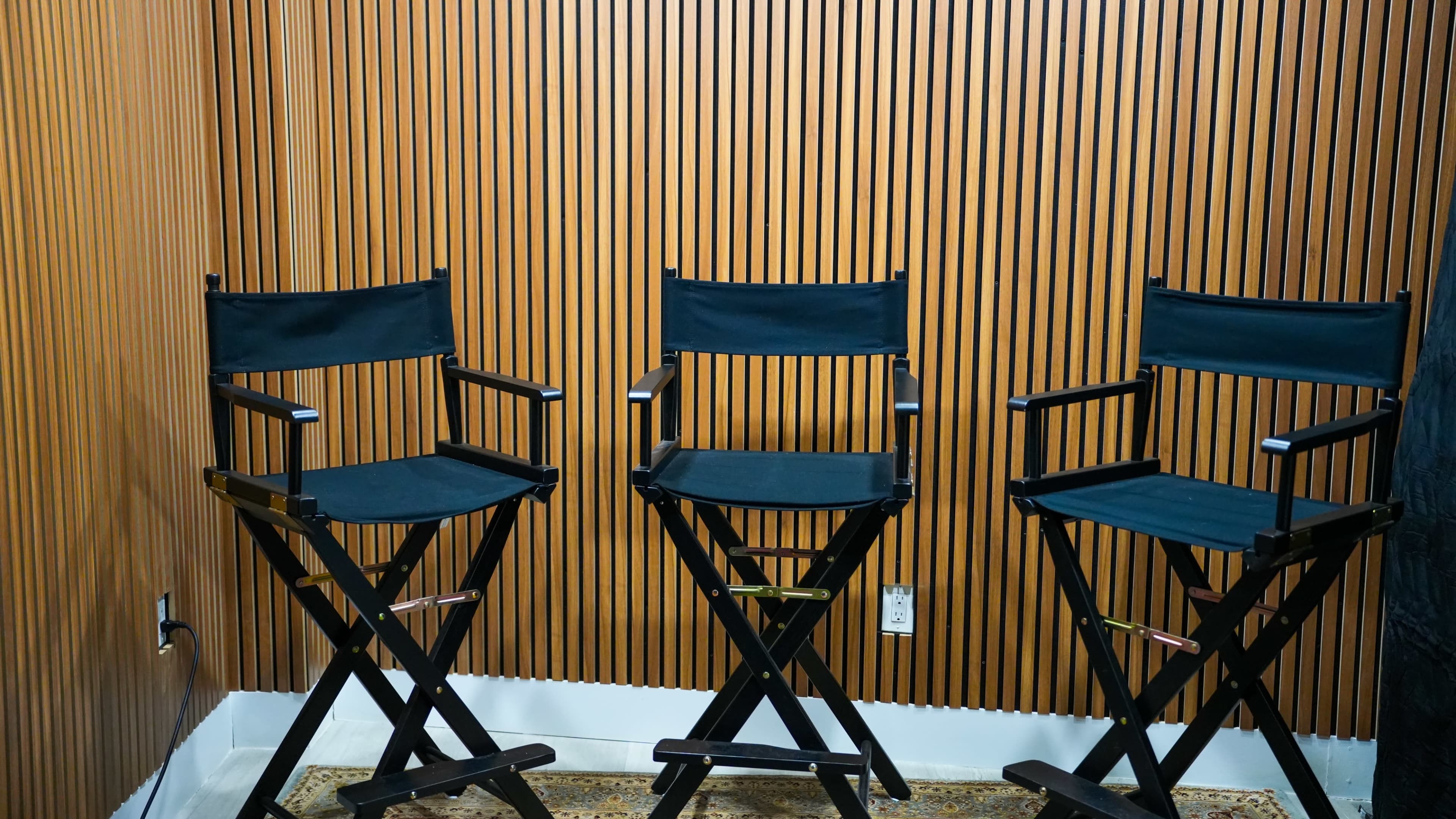 Three black director's chairs are arranged against a wooden slatted wall in a well-lit interior space.