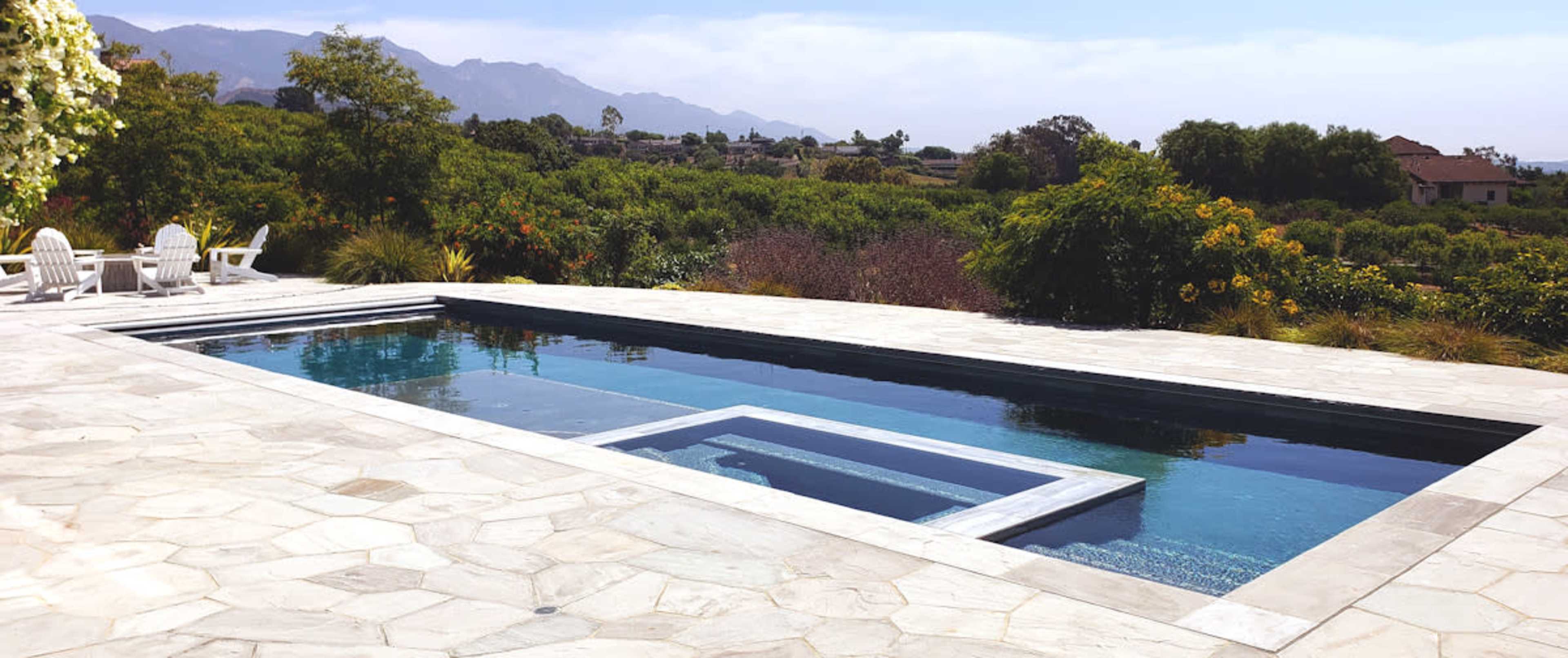 A rectangular swimming pool surrounded by stone patio, with lounge chairs positioned nearby and lush greenery in the background.