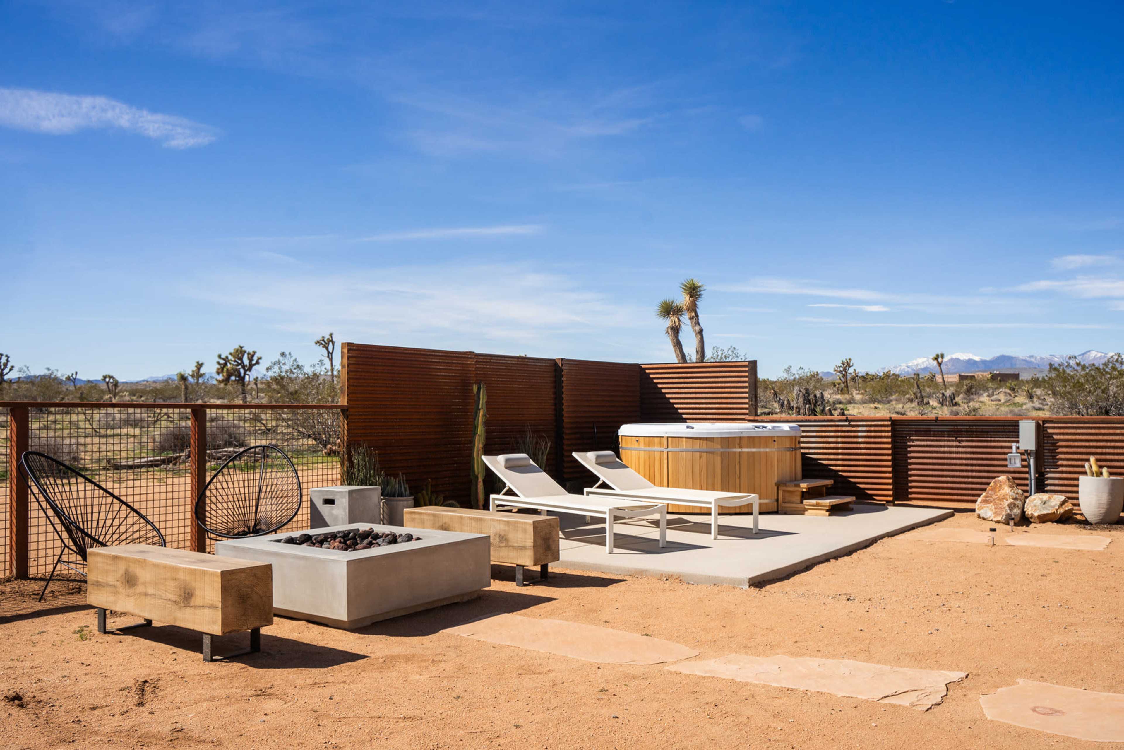 A minimalist outdoor area featuring a hot tub, two lounge chairs, and a fire pit surrounded by desert landscaping.