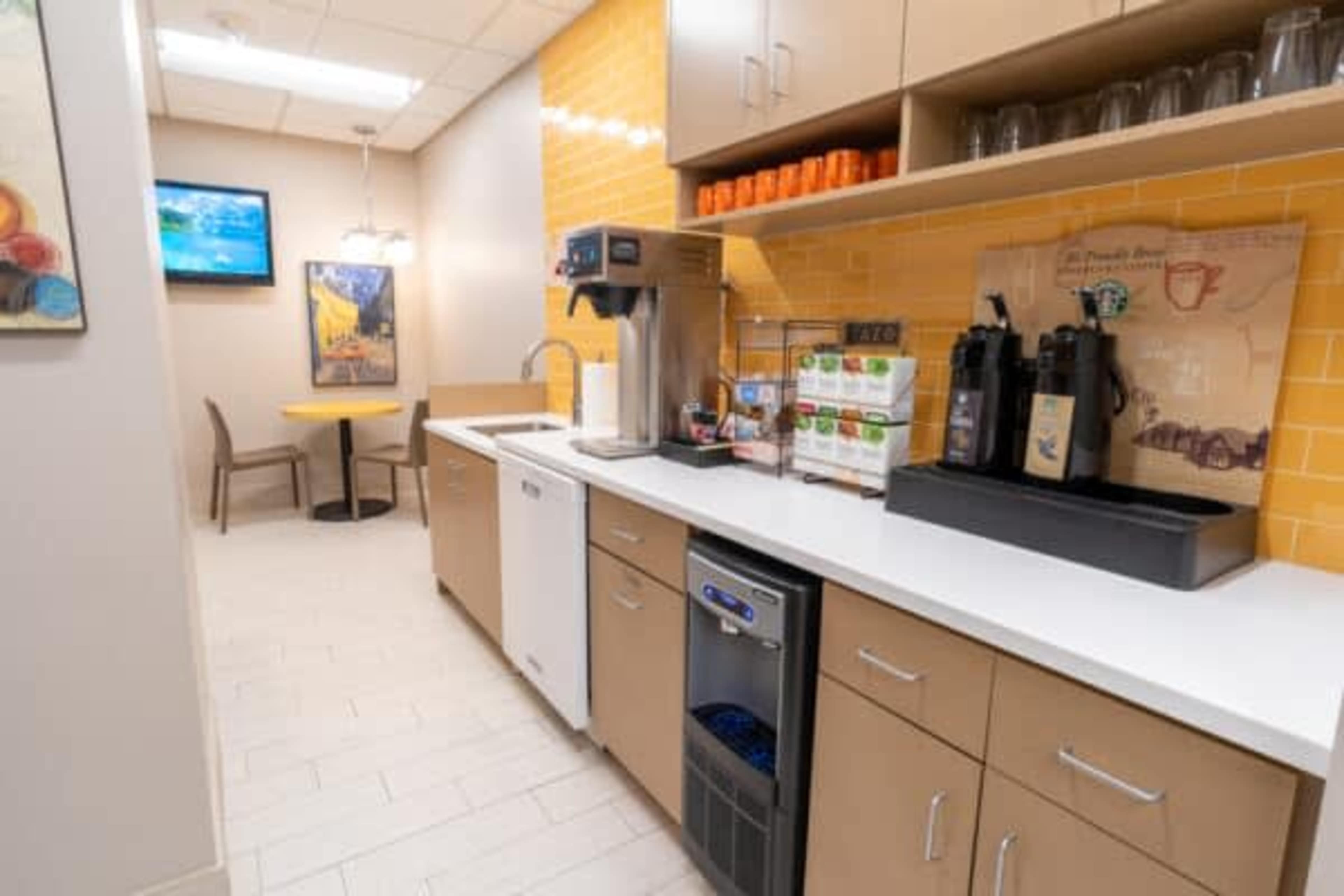 A modern break room features a coffee station, a dishwasher, and a small table with chairs under warm yellow tiled walls.