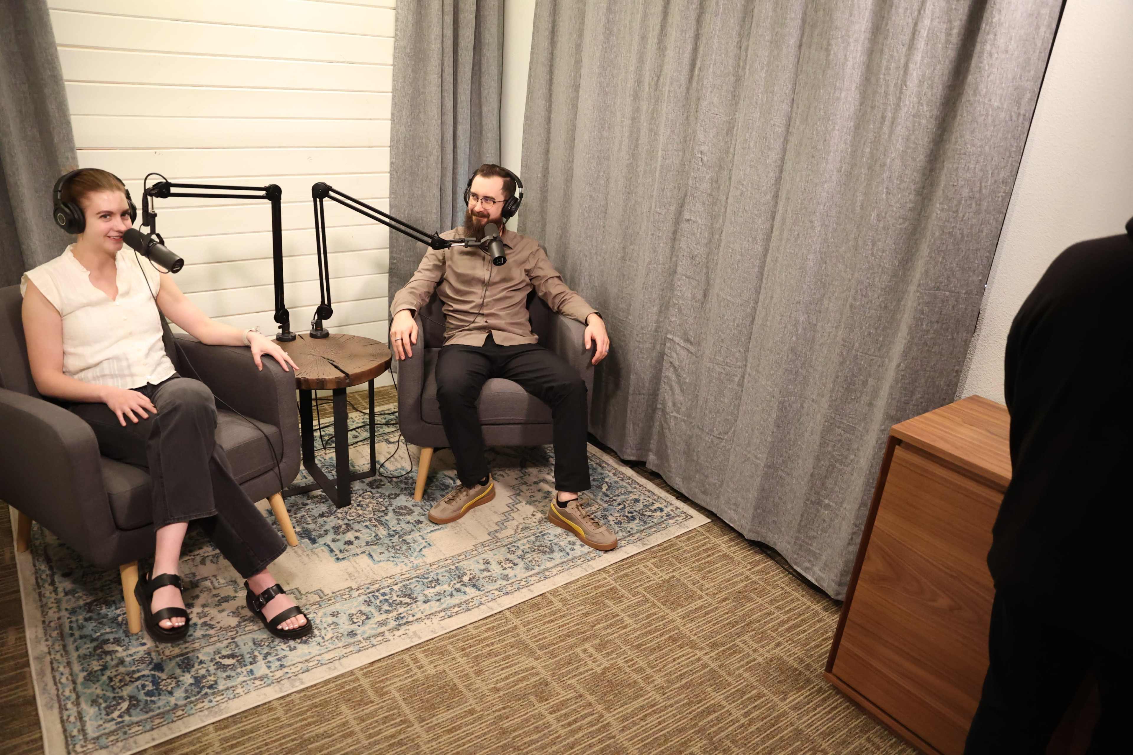 Two people sit in armchairs in a soundproof room equipped for podcasting, with microphones positioned in front of them.