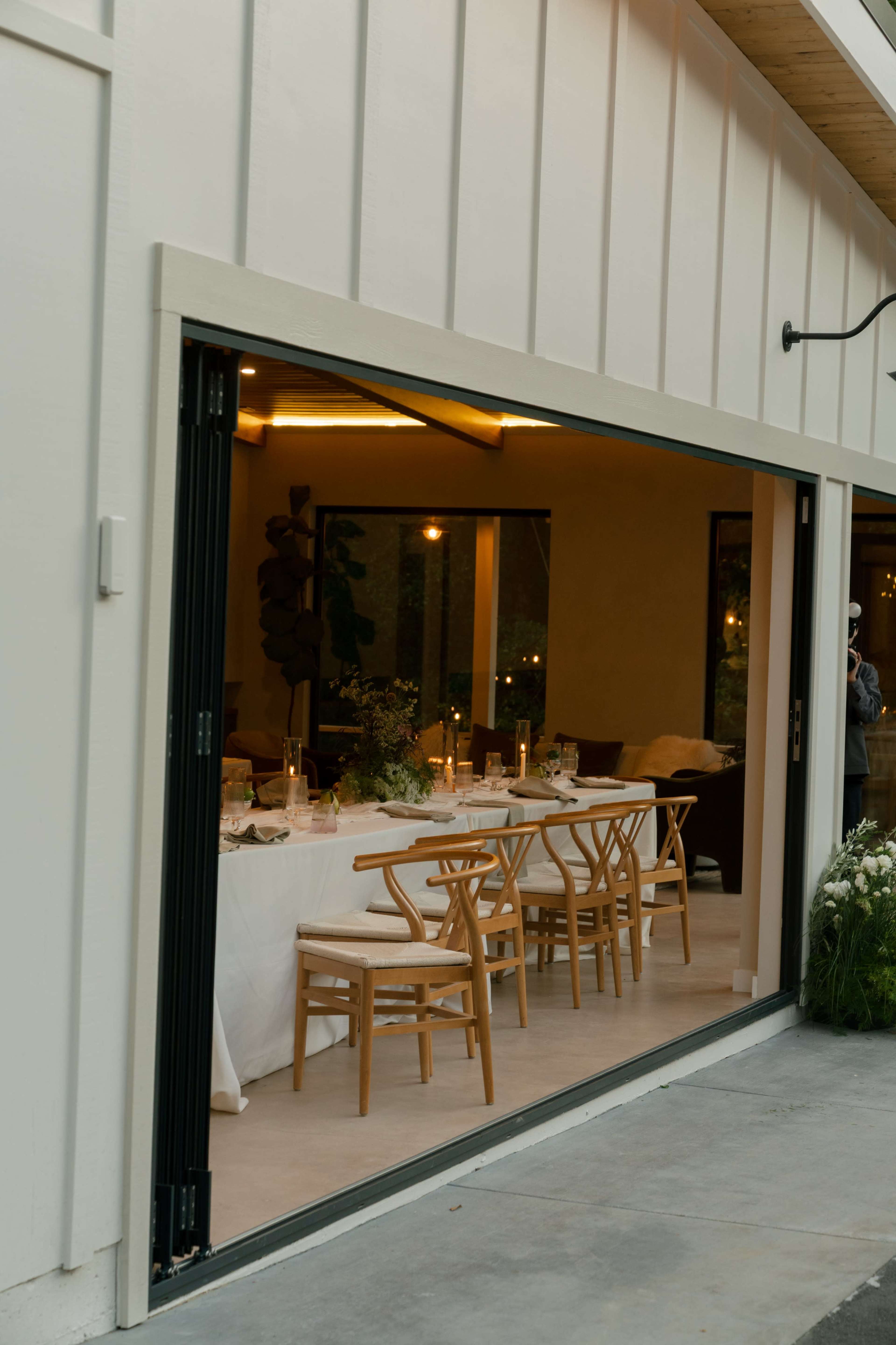 A dining area featuring a long table set with candles and floral arrangements is visible through large open doors.