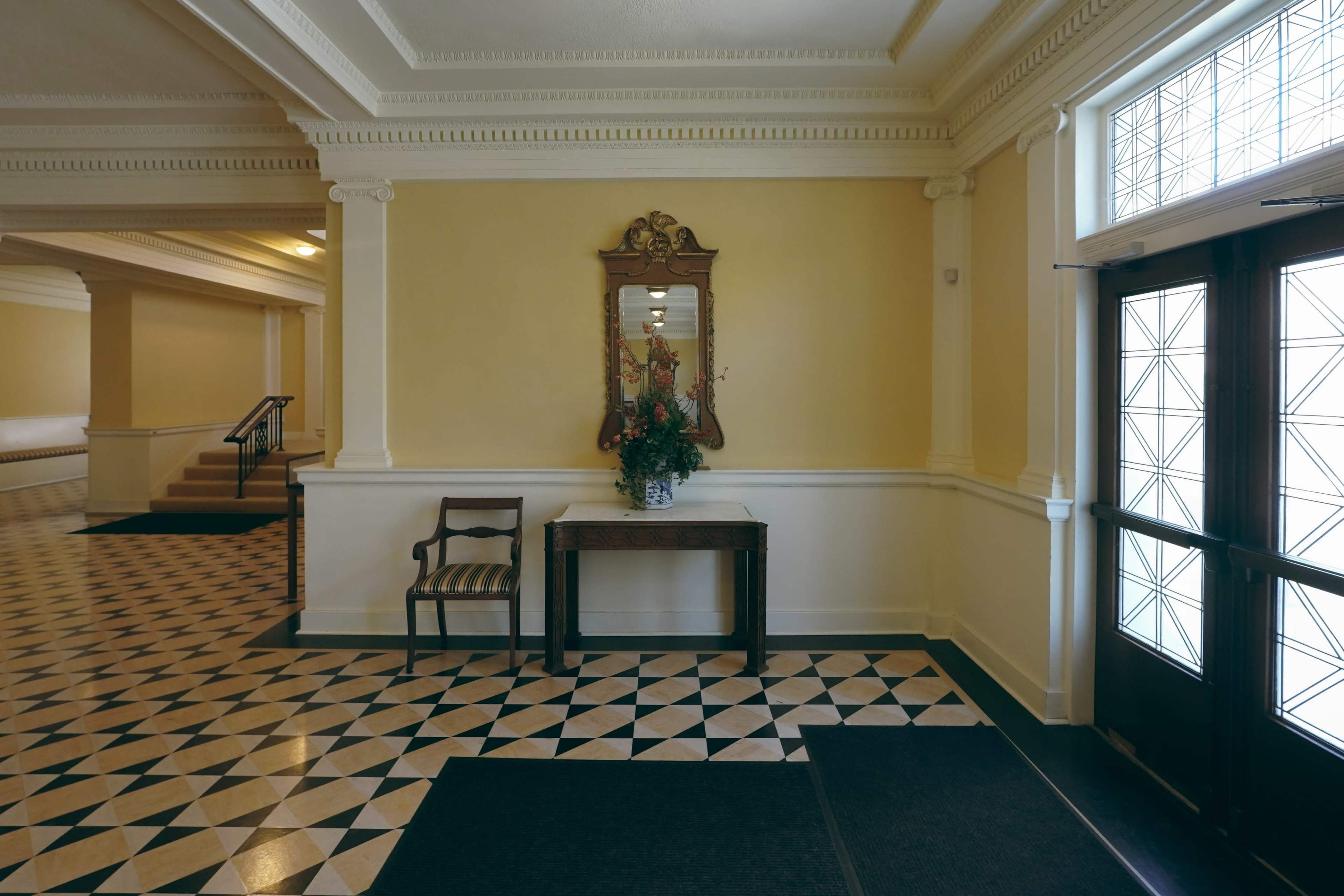 A spacious entryway with a patterned tiled floor, a wooden table with a vase of flowers, a mirror on the wall, and two doors leading outside.