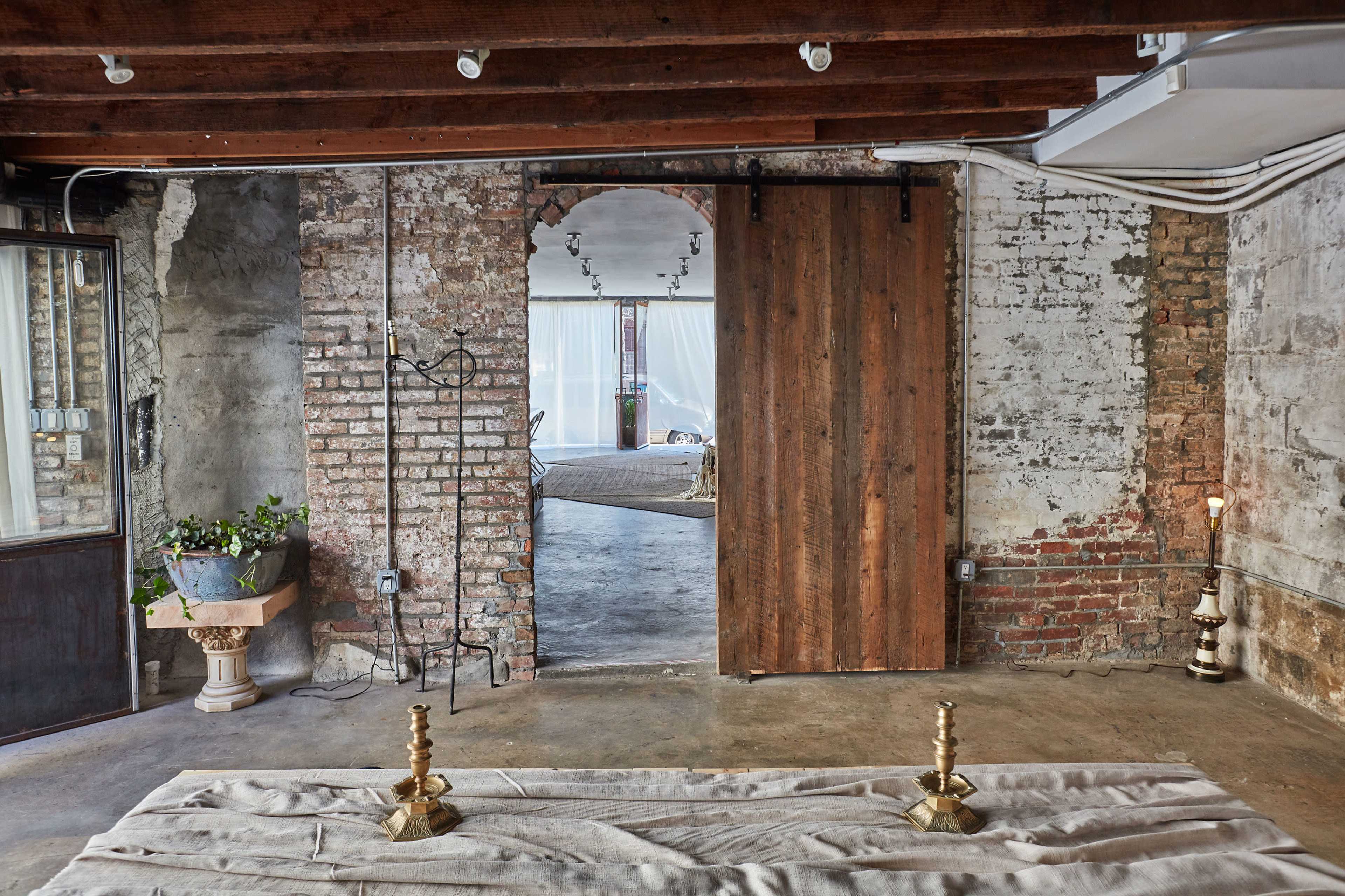 An interior space with exposed brick walls, wooden beams, and a doorway leading to another room, with decorative elements like candles and a potted plant on the floor.