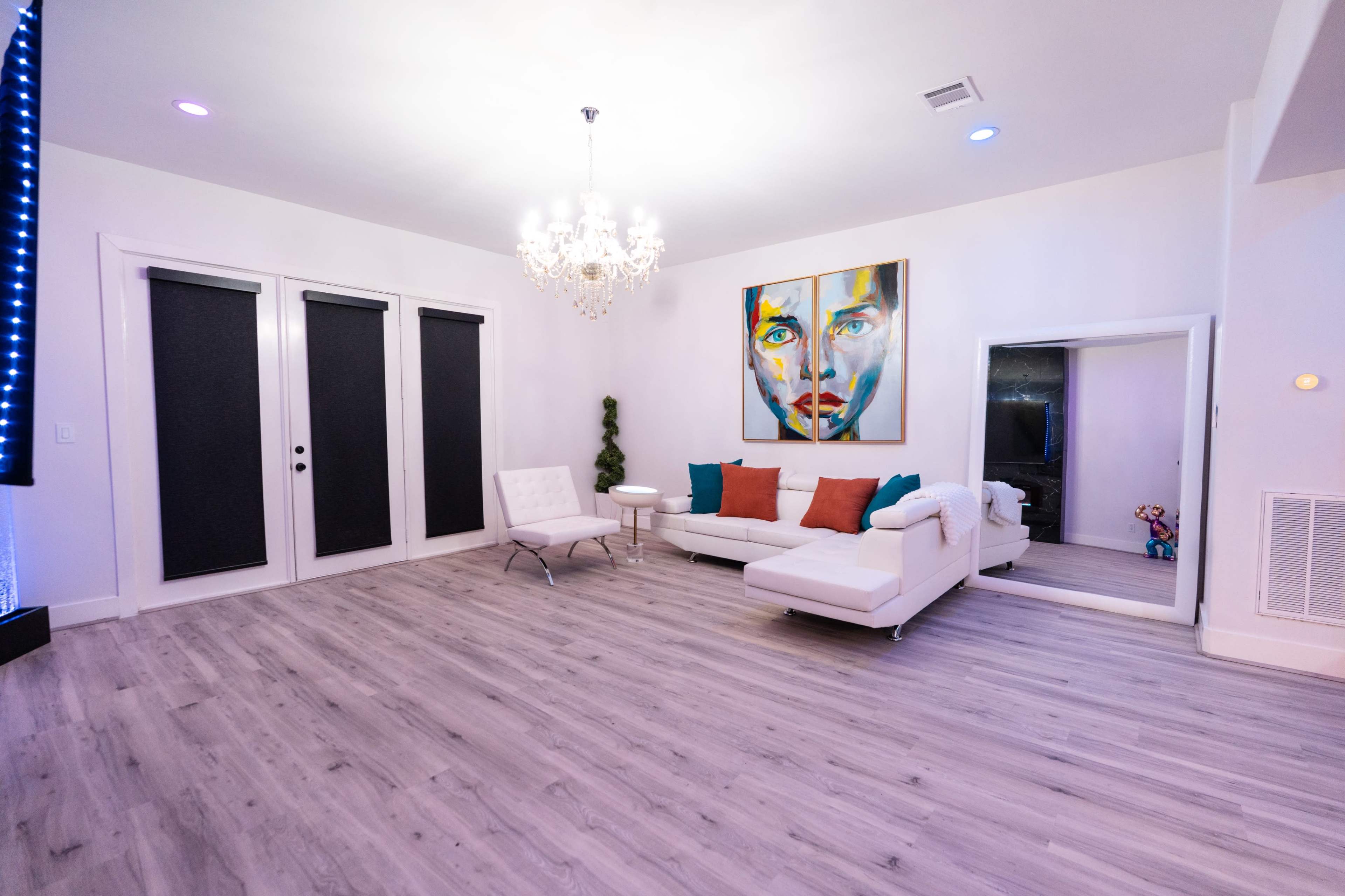The image shows a modern living room featuring a white sectional sofa, a large colorful portrait on the wall, a chandelier, and a mirror, with light-colored flooring.