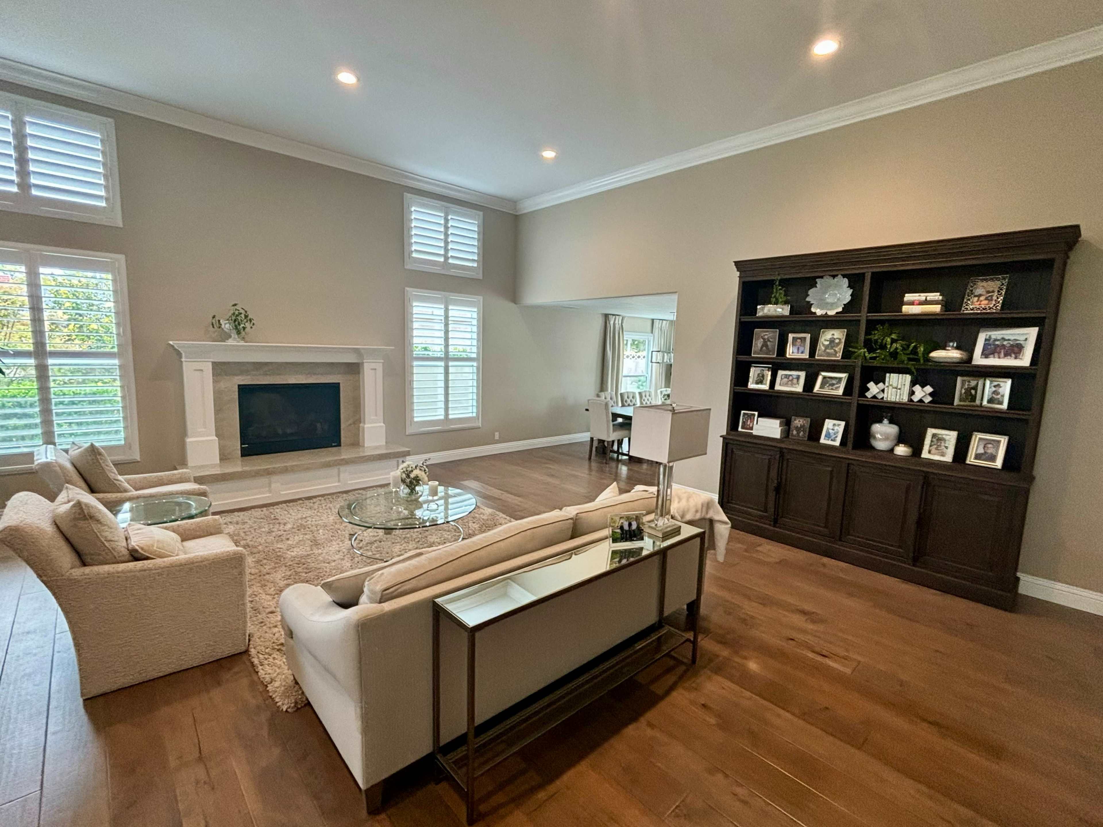 A spacious living room features a fireplace, large windows, and a dark wooden bookshelf filled with framed photos and decorative items.