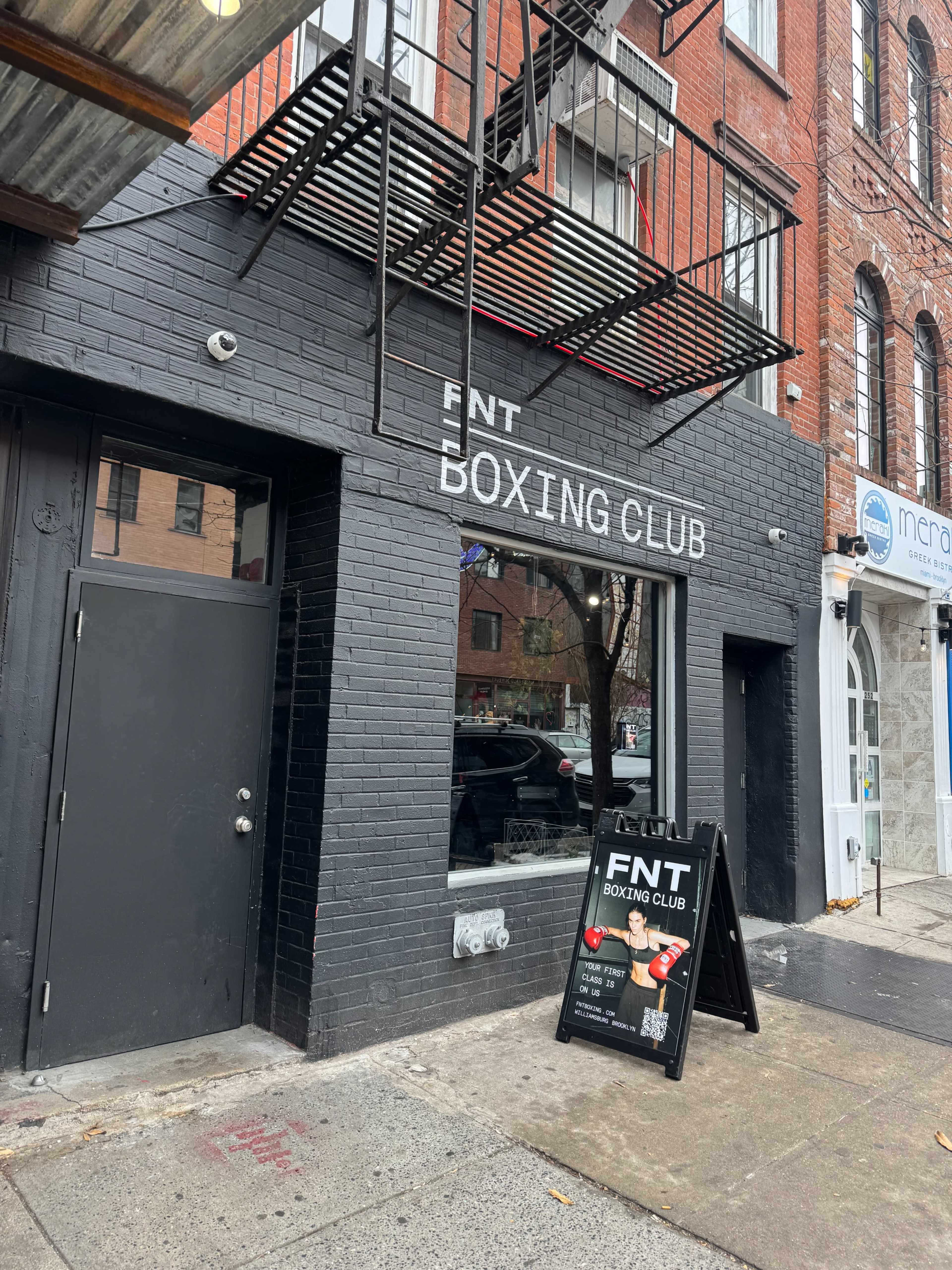 The image shows the exterior of a boxing club named "FNT Boxing Club," with a sign displaying its name and a promotional poster on an A-frame board outside.