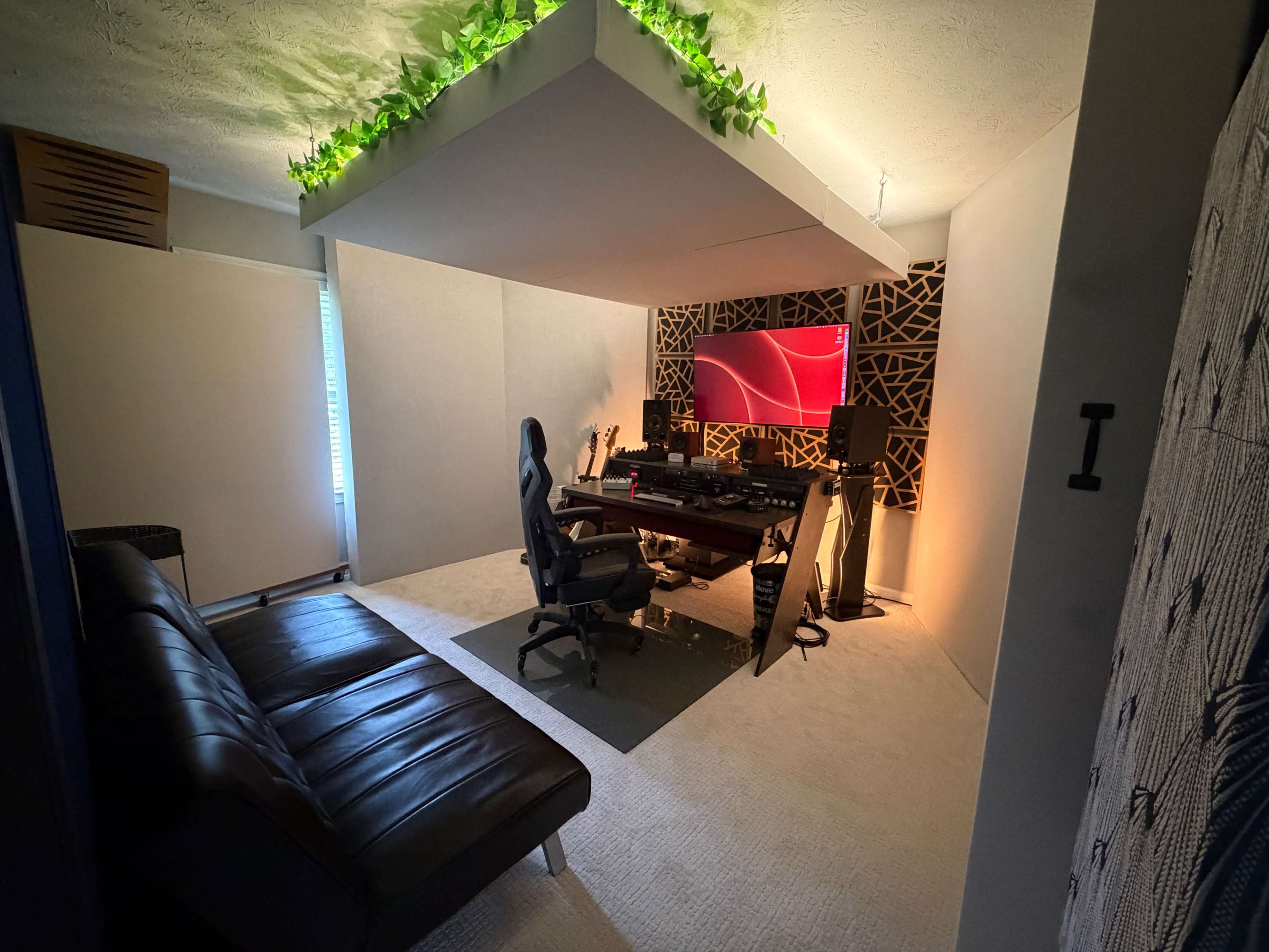 The image shows a modern room equipped with a gaming setup, featuring a desk with multiple monitors, a black gaming chair, a couch, and decorative lighting.