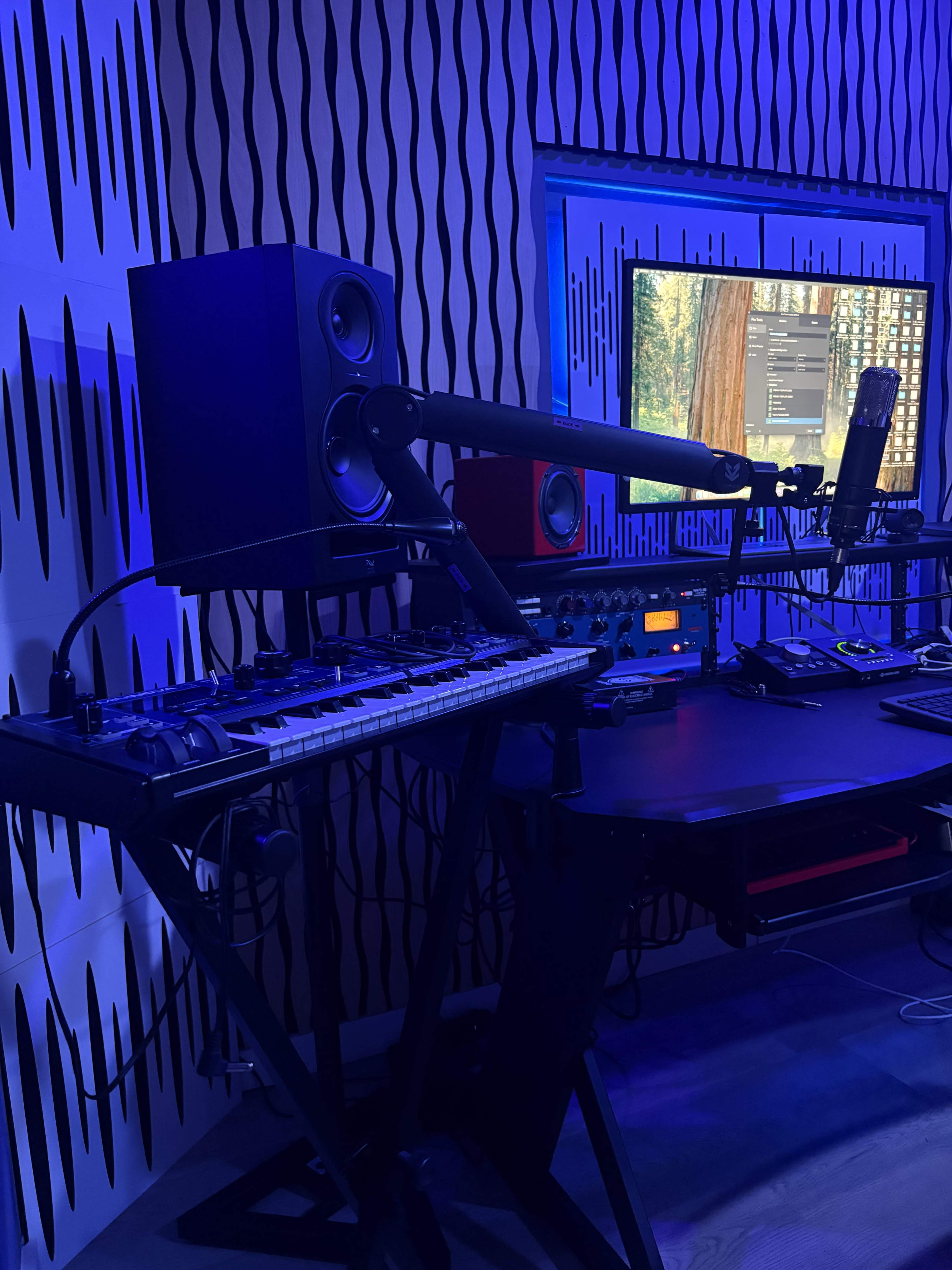 The image shows a music production setup featuring a keyboard, monitors, a microphone, and a computer screen, all set against a textured wall with blue lighting.